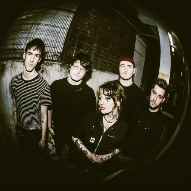 Group of five young adults standing outdoors at night, with a brick wall and metal fence behind them.