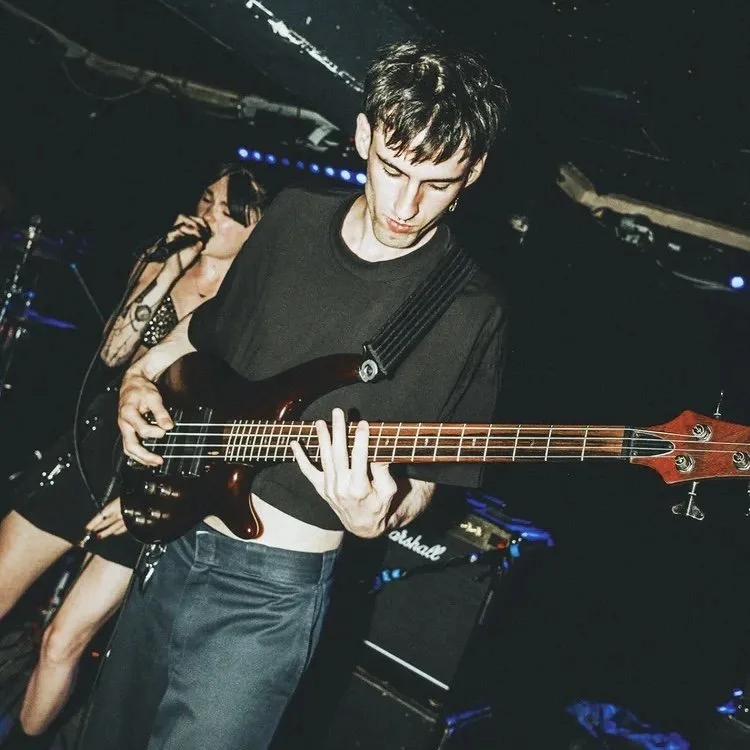 A young man with dark hair wearing a black T-shirt playing a bass guitar on stage during a live performance, with a female vocalist singing into a microphone in the background.