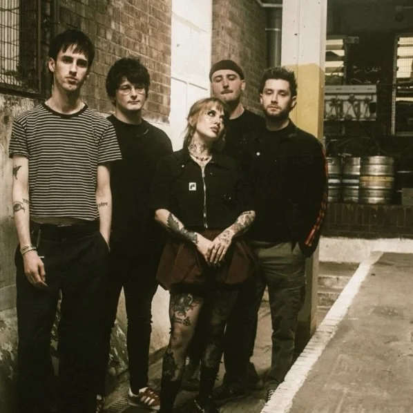 Group of five young adults standing outdoors near a brick wall and parked kegs, with a brick and sidewalk background.