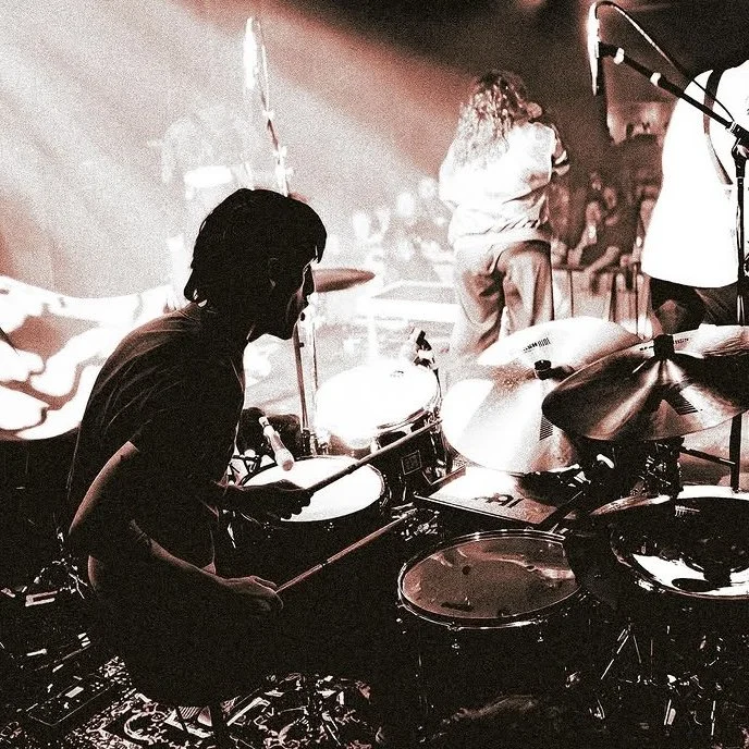 A drummer playing drums on stage during a live music performance with other musicians and an audience in the background.
