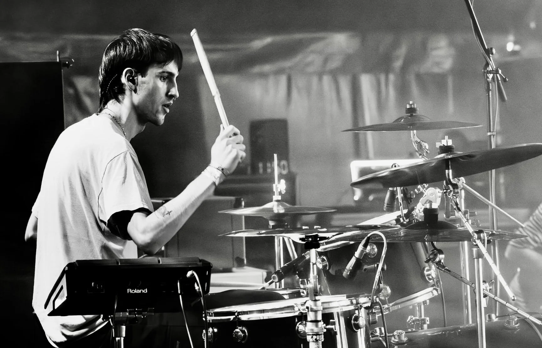 A young man with dark hair playing drums on a stage, wearing a light-colored t-shirt, surrounded by drum set components and electronic equipment, in black and white.