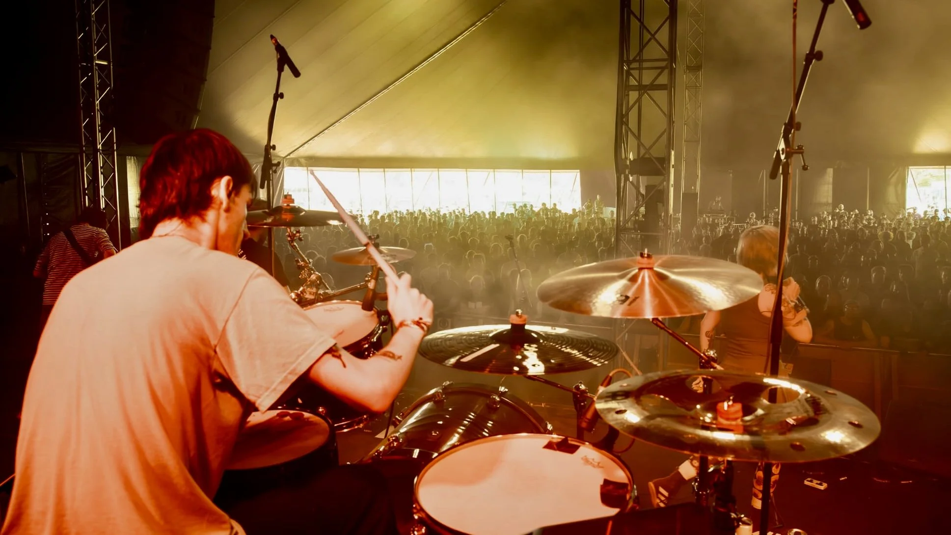Photograph of a live music performance from behind the drums, with audience in the background inside a large tent or pavilion.
