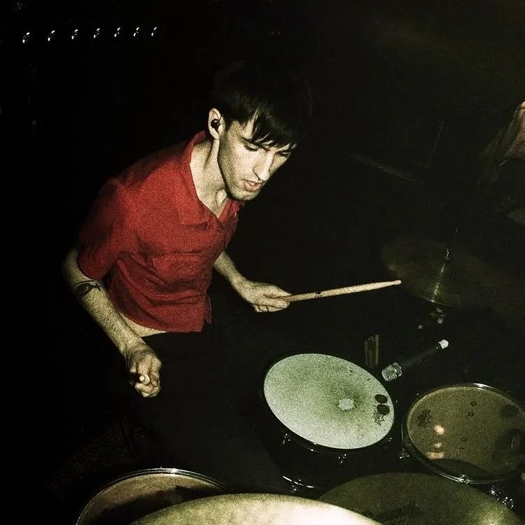 A man playing drums in a dark room, wearing a red shirt and holding drumsticks.