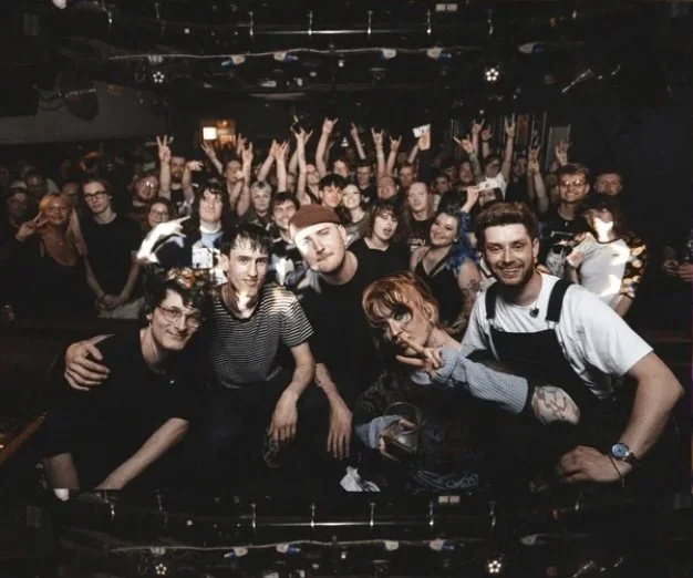 Crowd of people at a concert or event, with five individuals in the front posing for the photo, some making peace signs and smiling.