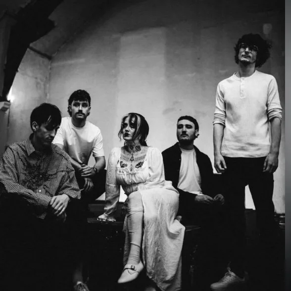 Black and white photo of five people sitting and standing against a plain wall, with one woman in a long dress and four men in casual clothing.