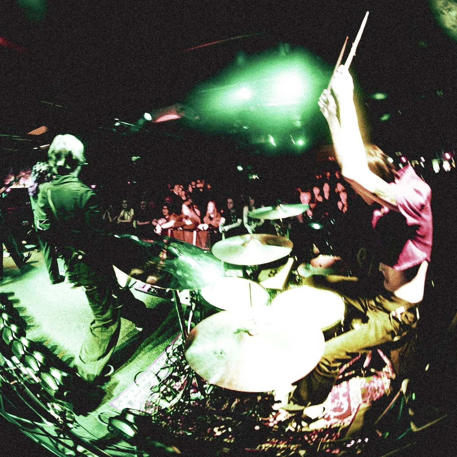 A drummer with a pink shirt playing drums on stage while an audience watches at a concert with green stage lighting.