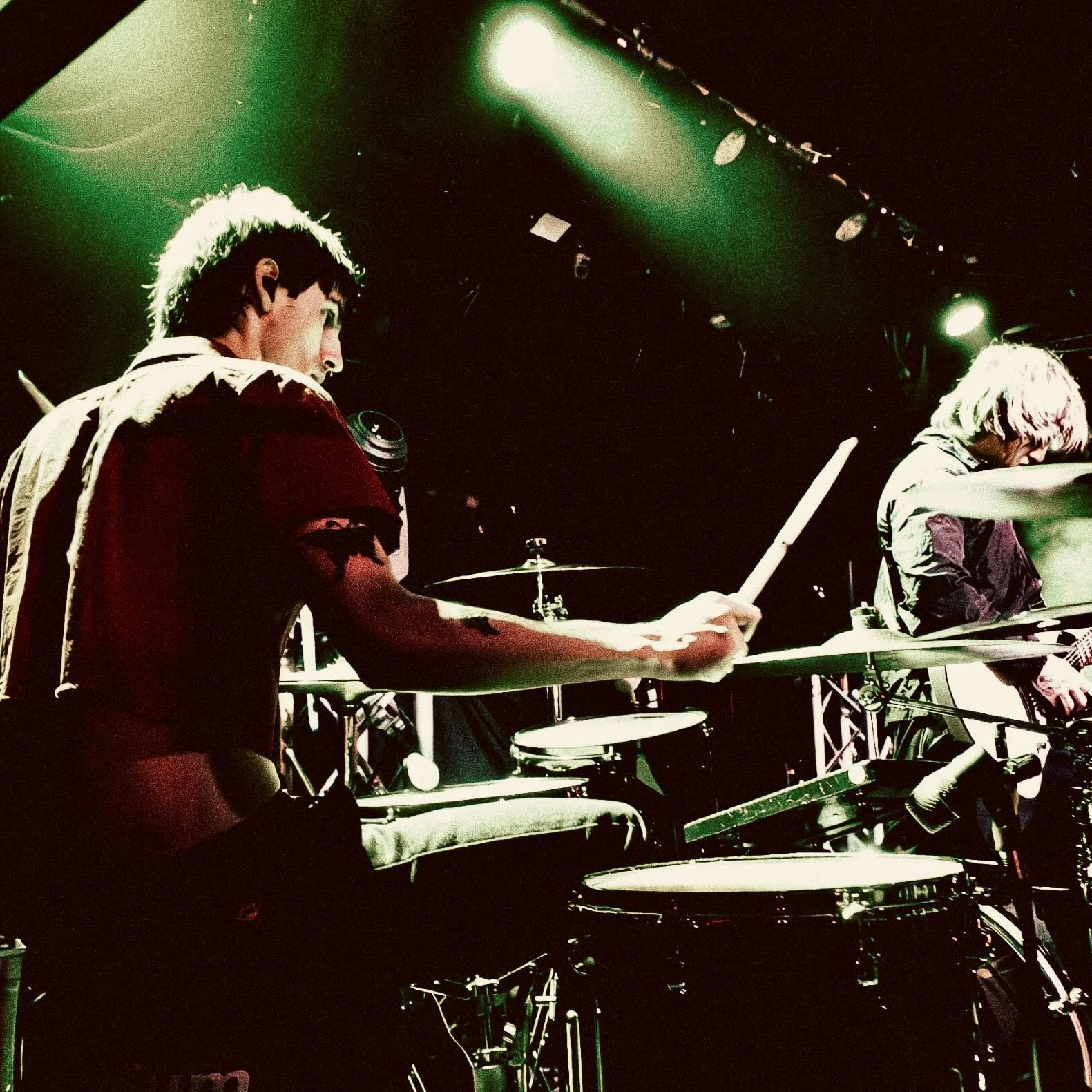 Two musicians performing on stage, playing drums and a keyboard, with stage lighting creating green and white highlights.