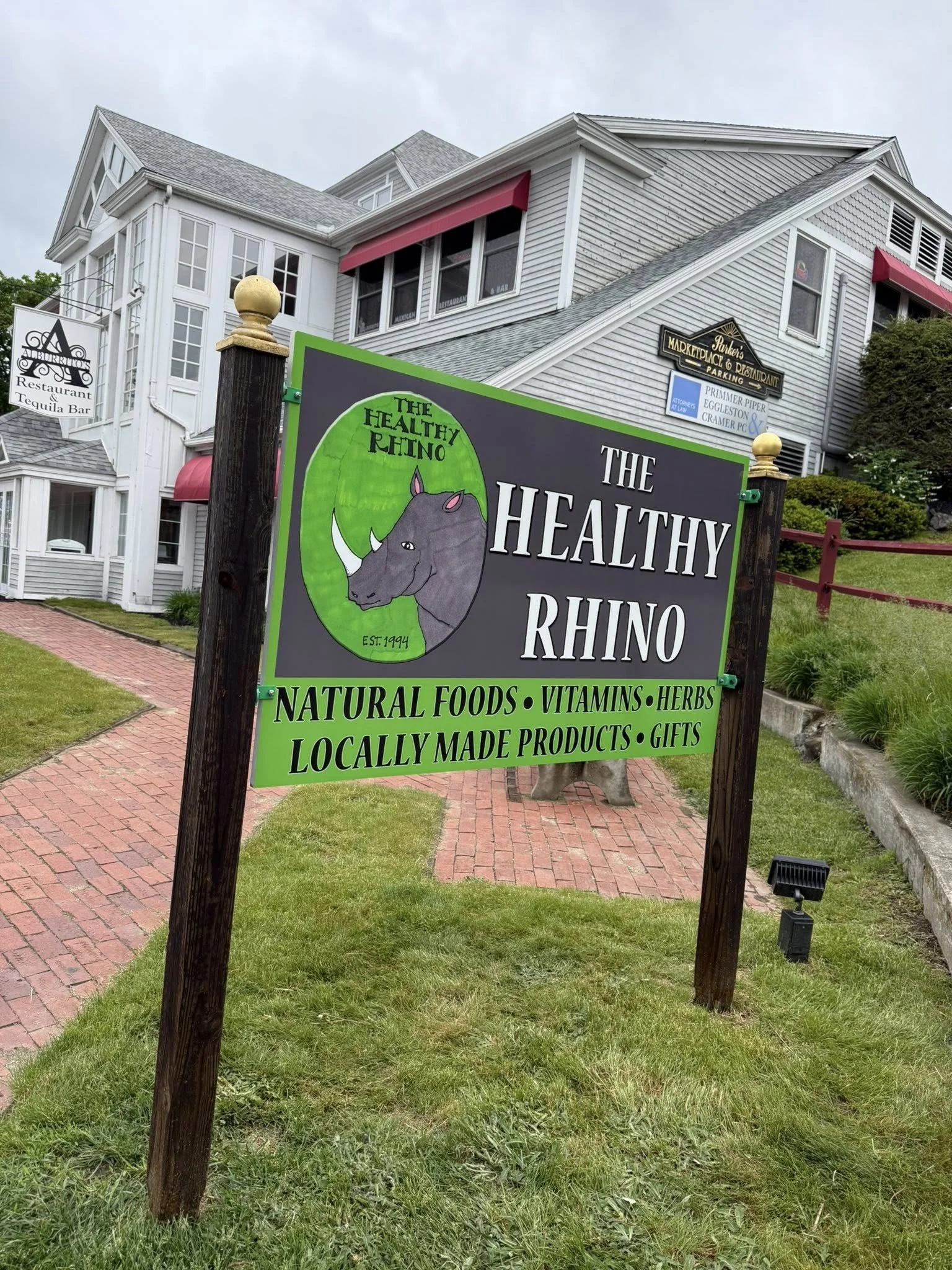 Sign for The Healthy Rhino, a business selling natural foods, vitamins, herbs, locally made products, and gifts, with a drawing of a rhinoceros on the sign. The background shows a large building with multiple windows, a parking lot, and signs for a restaurant and other businesses.