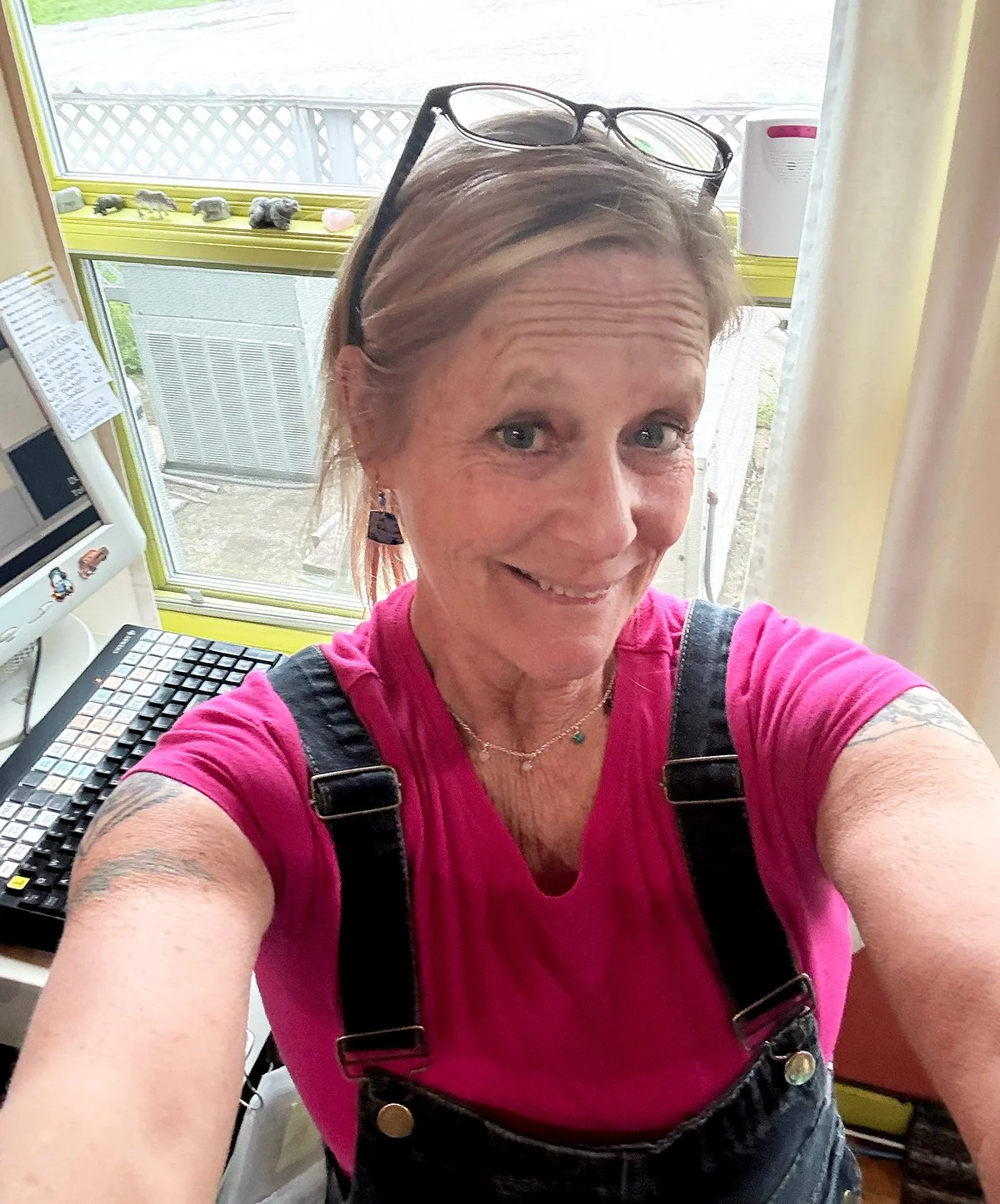 A woman taking a selfie inside a room near a window, wearing a pink shirt, black overalls, and glasses on her head, with a computer keyboard and monitor behind her.