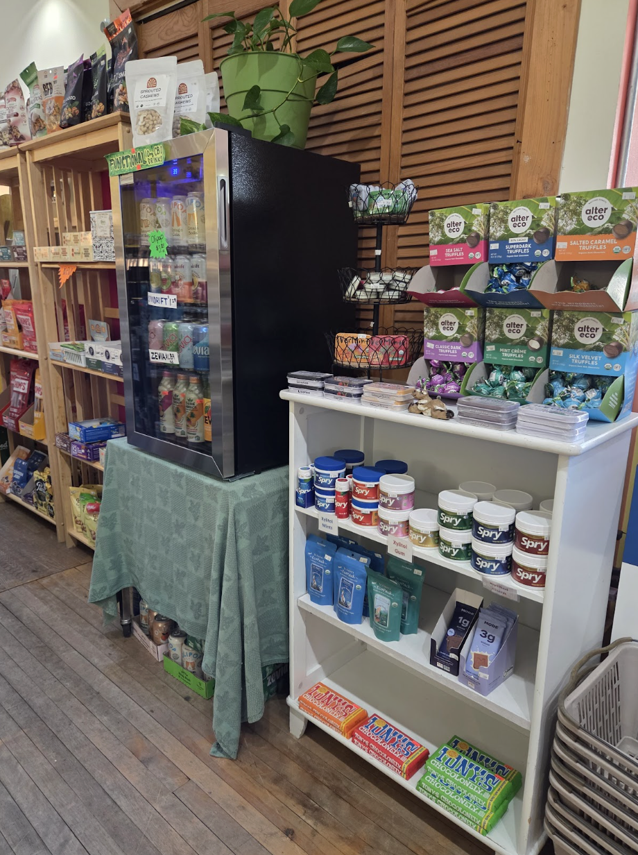 A display of assorted chocolates, supplements, and health products in a store with wooden shelves and flooring. The display includes a mini fridge, truffle boxes, and containers of Spry mints.