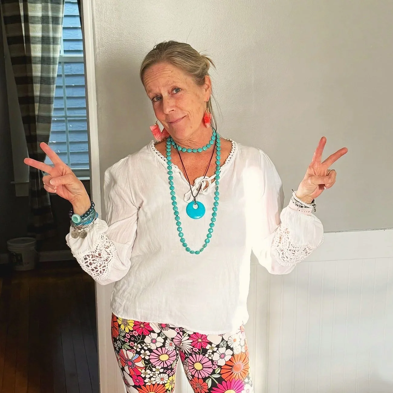 Woman posing with peace signs, wearing a white blouse with lace sleeves, floral patterned pants, and colorful jewelry, standing in a room with a window and curtains in the background.