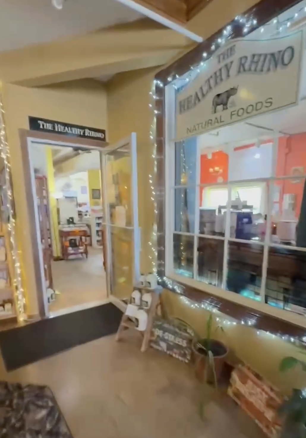 Entrance to a store called The Healthy Rhino, decorated with string lights, with a window displaying the interior and a door to the left leading inside. There is a black mat on the floor and some potted plants and signs near the window.