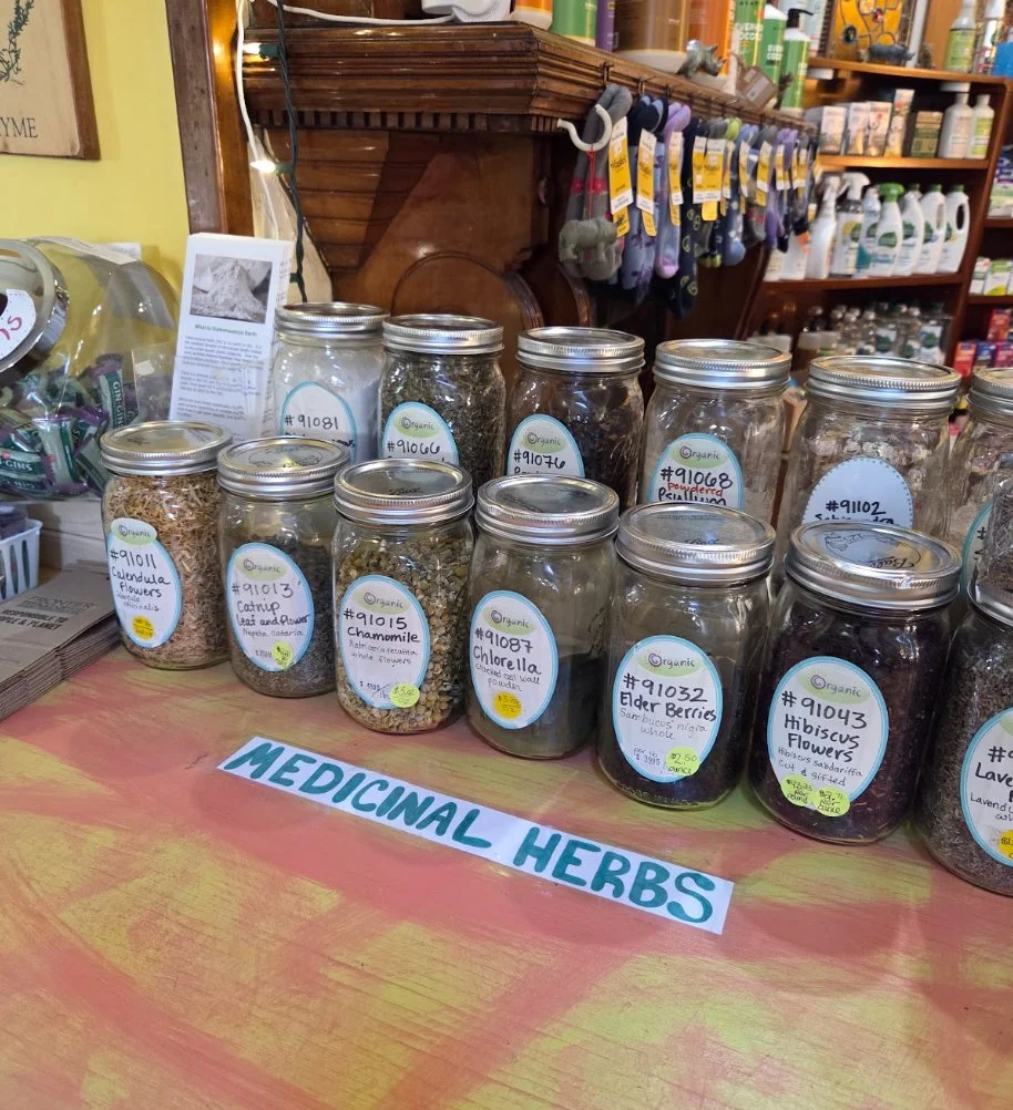Collection of glass jars filled with various dried herbs labeled as organic medicinal herbs, displayed on a table with a sign reading 'MEDICINAL HERBS' in a store.