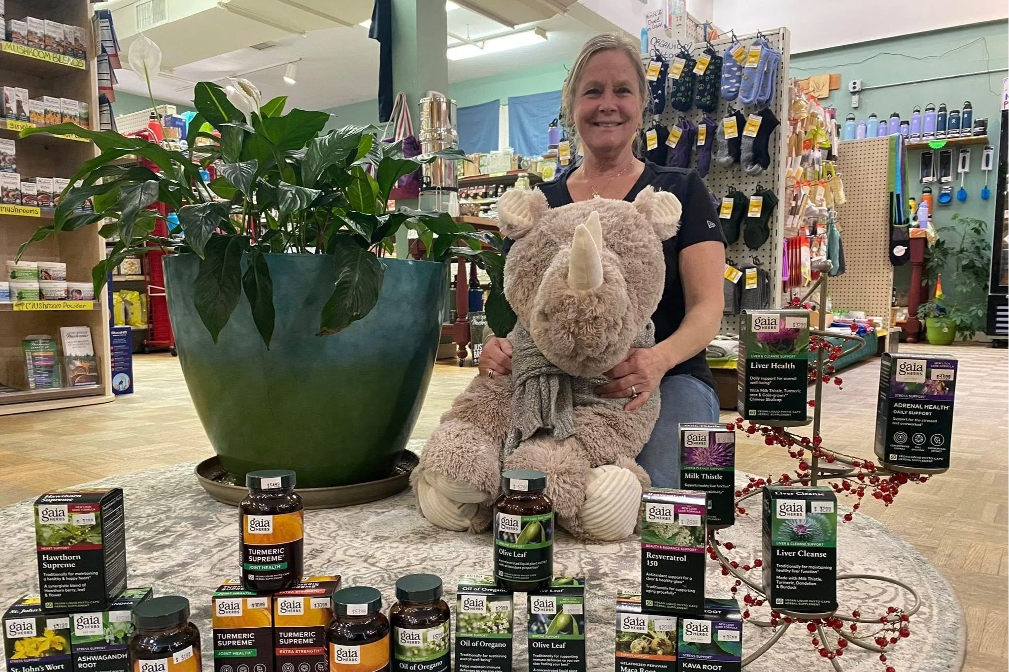 A woman smiling and holding a large plush stuffed bear with a unicorn horn in a store. There are shelves with various products in the background and a large potted plant in the foreground.