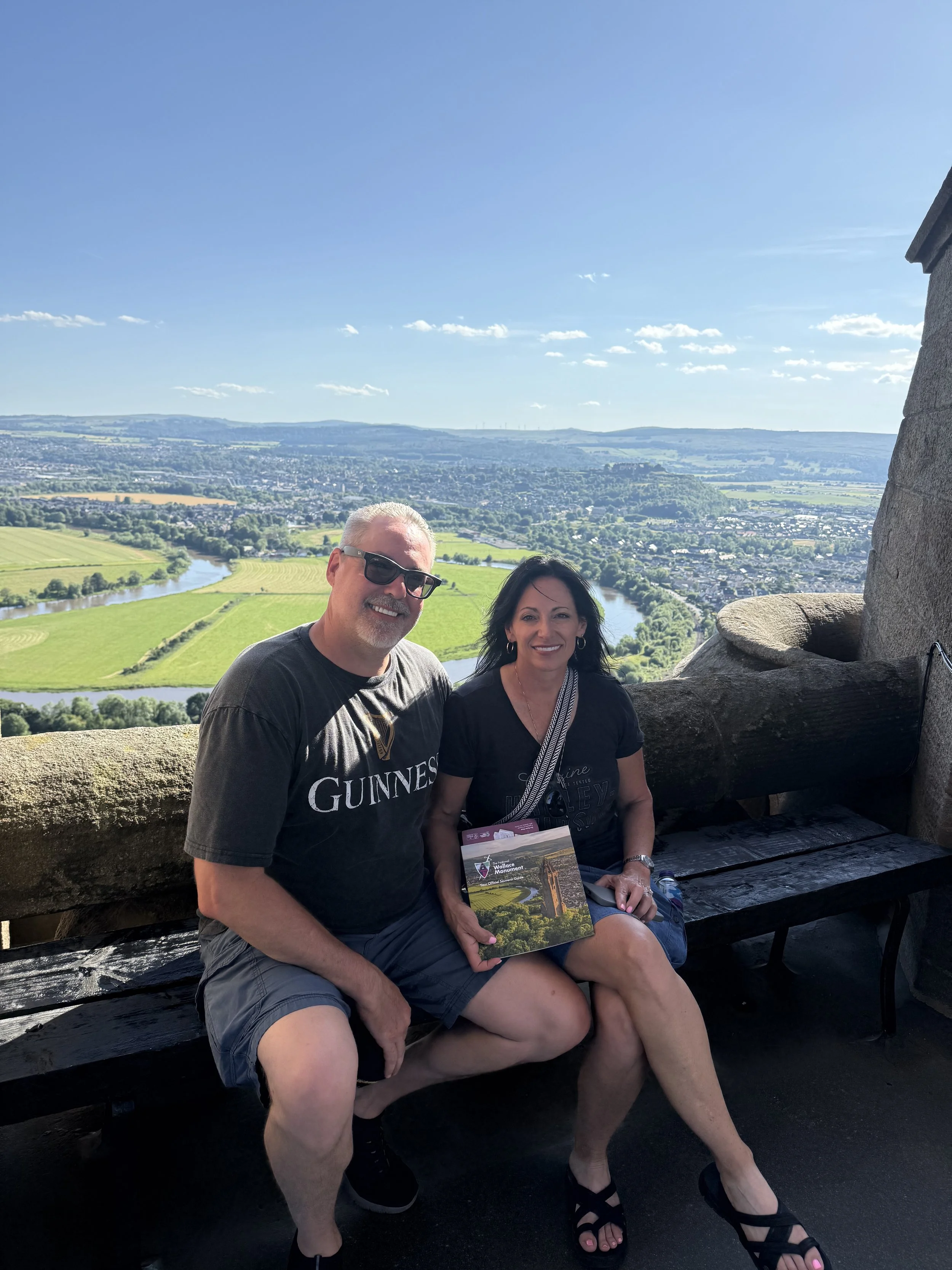 A smiling man and woman sitting on a wooden bench overlooking a scenic landscape with river, fields, and a distant town under a clear blue sky. The man wears sunglasses and a black Guinness shirt, and the woman holds a pamphlet.