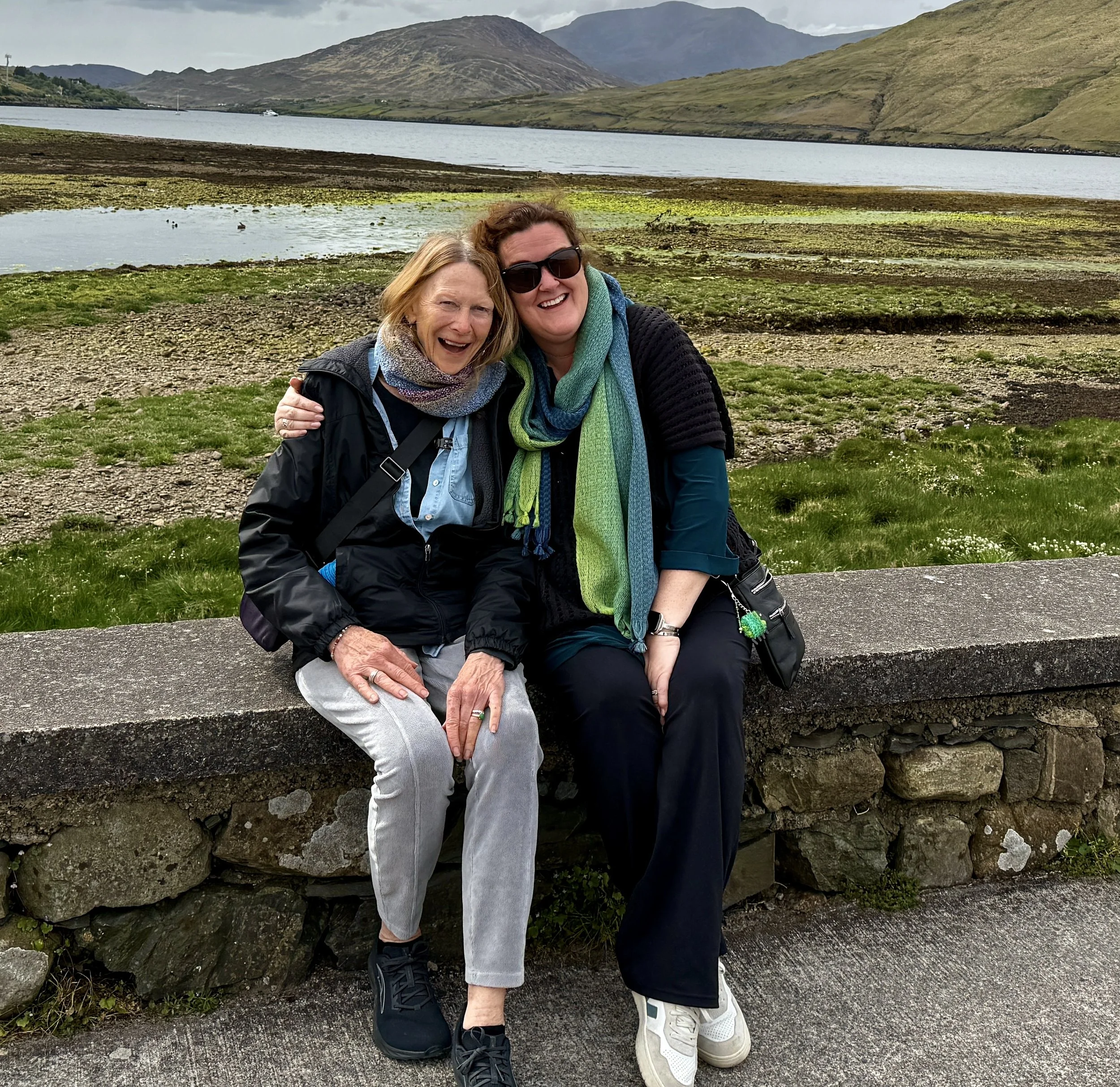 Two women sitting on a stone wall near a lake with mountains in the background. One woman has red hair and is wearing a black jacket, the other has brown hair and is wearing sunglasses and a black sweater. They are smiling and appear happy.