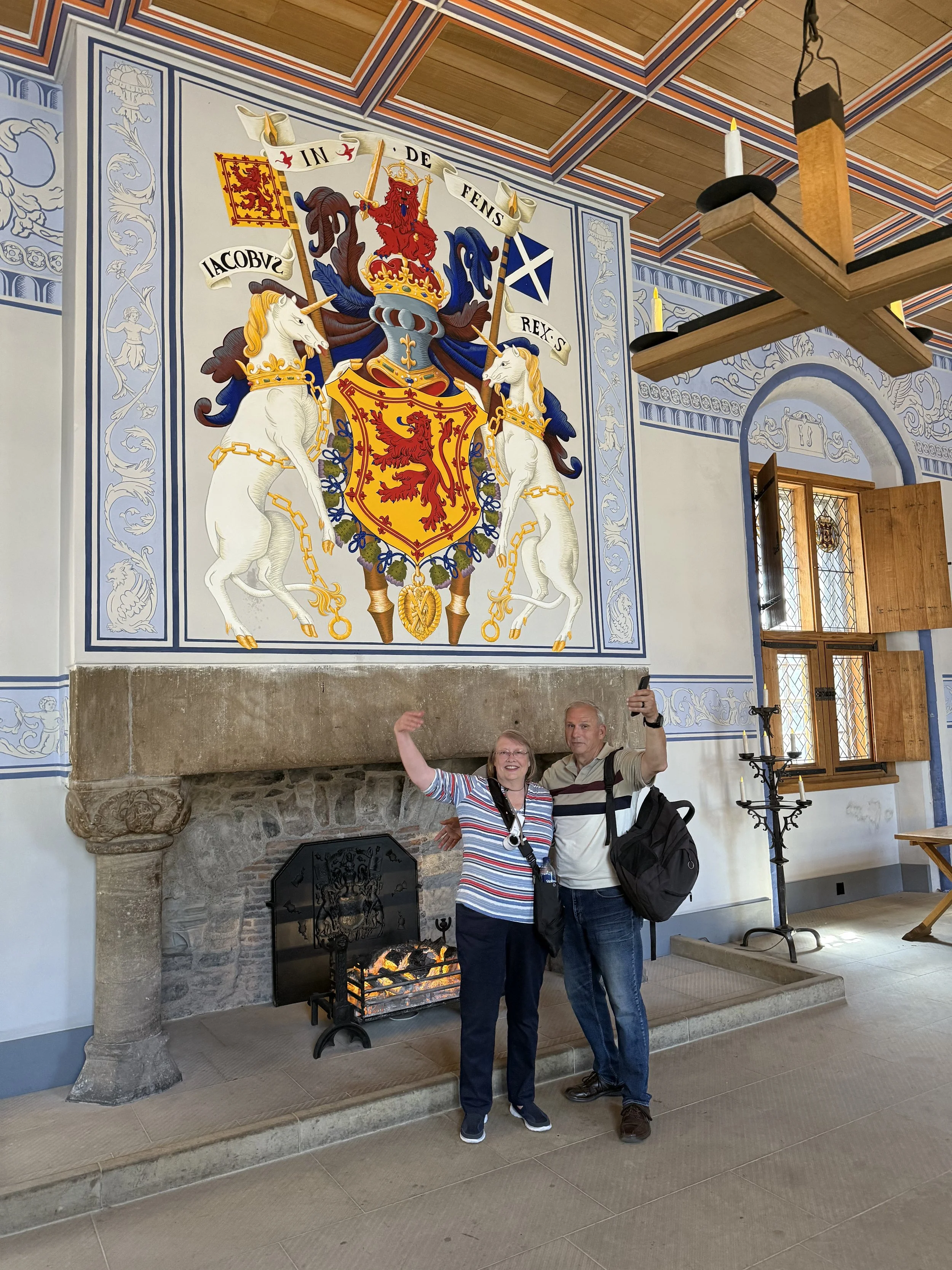 Two people, a woman and a man, smiling and posing for a selfie in front of a large coat of arms or crest on the wall of a historic building. The crest features a shield with a red lion and a crown, flanked by two white unicorns with gold chains. There is a fireplace with a fire burning below the crest and a wooden window with stained glass to the right.