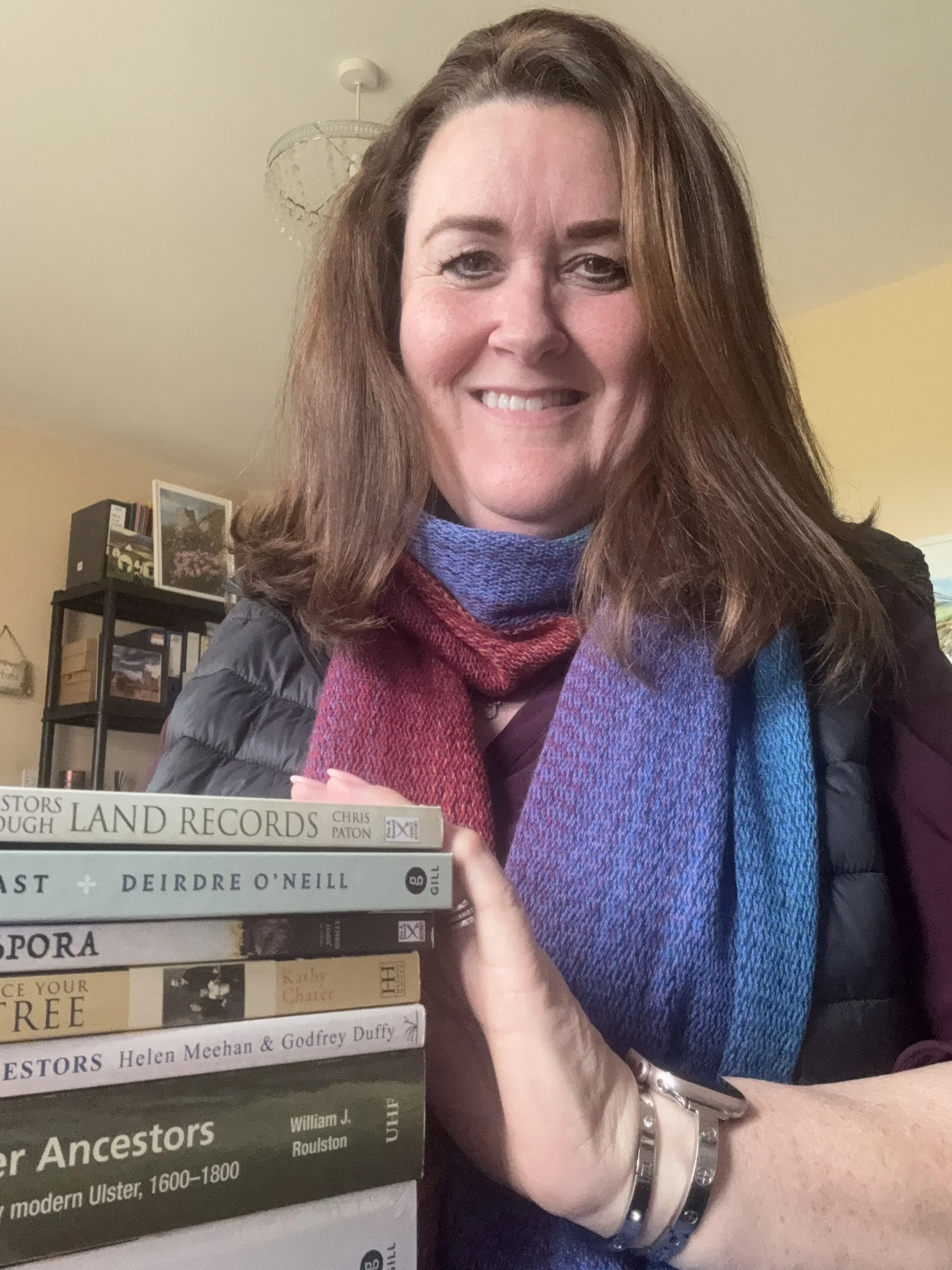 A woman with long brown hair smiling, wearing a blue and purple scarf, sitting indoors behind a stack of books on ancestors and history, with a bookshelf and framed picture in the background.