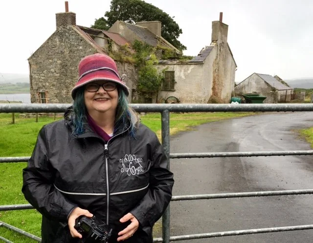 A woman wearing a pink hat, glasses, and a black jacket standing outdoors in front of an old stone building and a metal fence during cloudy weather.