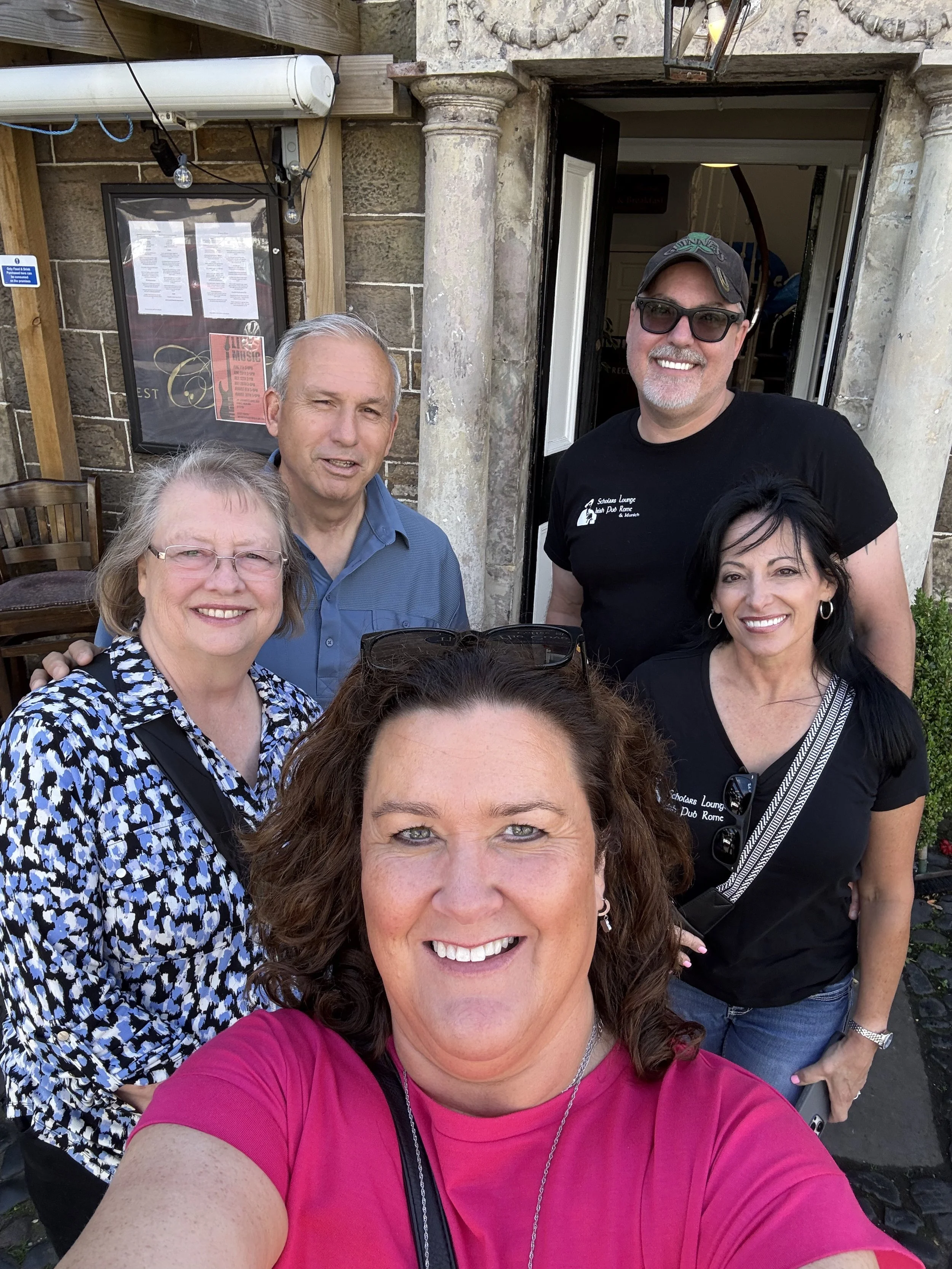 Group of five smiling people taking a selfie outside a building with stone and brick walls. The woman in front has curly brown hair and is wearing a pink shirt. Behind her, an older woman with glasses and a patterned blouse, a man with gray hair in a blue shirt, a man with a beard, sunglasses, and a cap, and a woman with dark hair and earrings are visible.