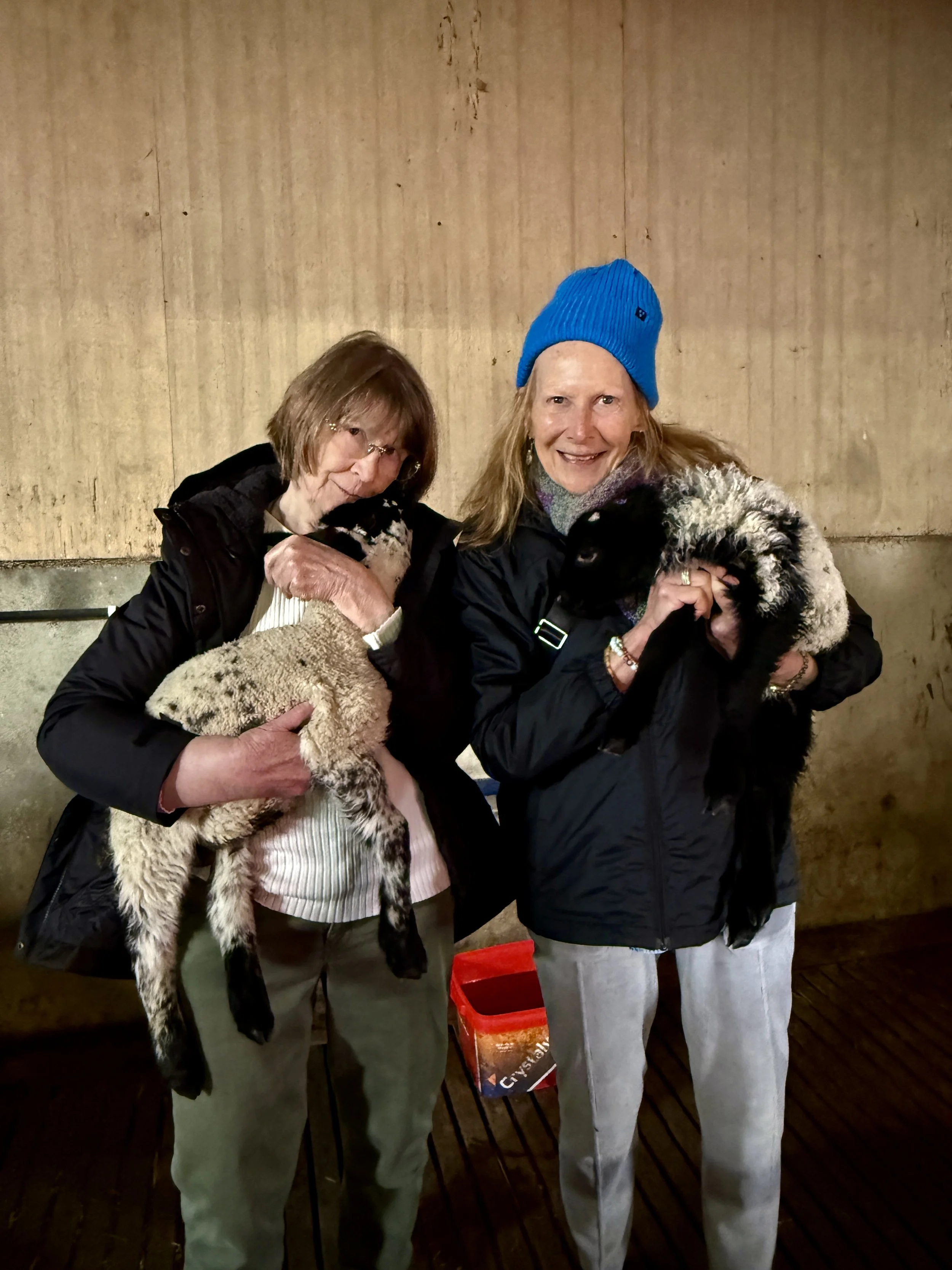 Two women holding puppies, standing in front of a wooden wall. The woman on the left has glasses and shoulder-length hair, while the woman on the right wears a blue beanie and has long hair. Both are smiling.