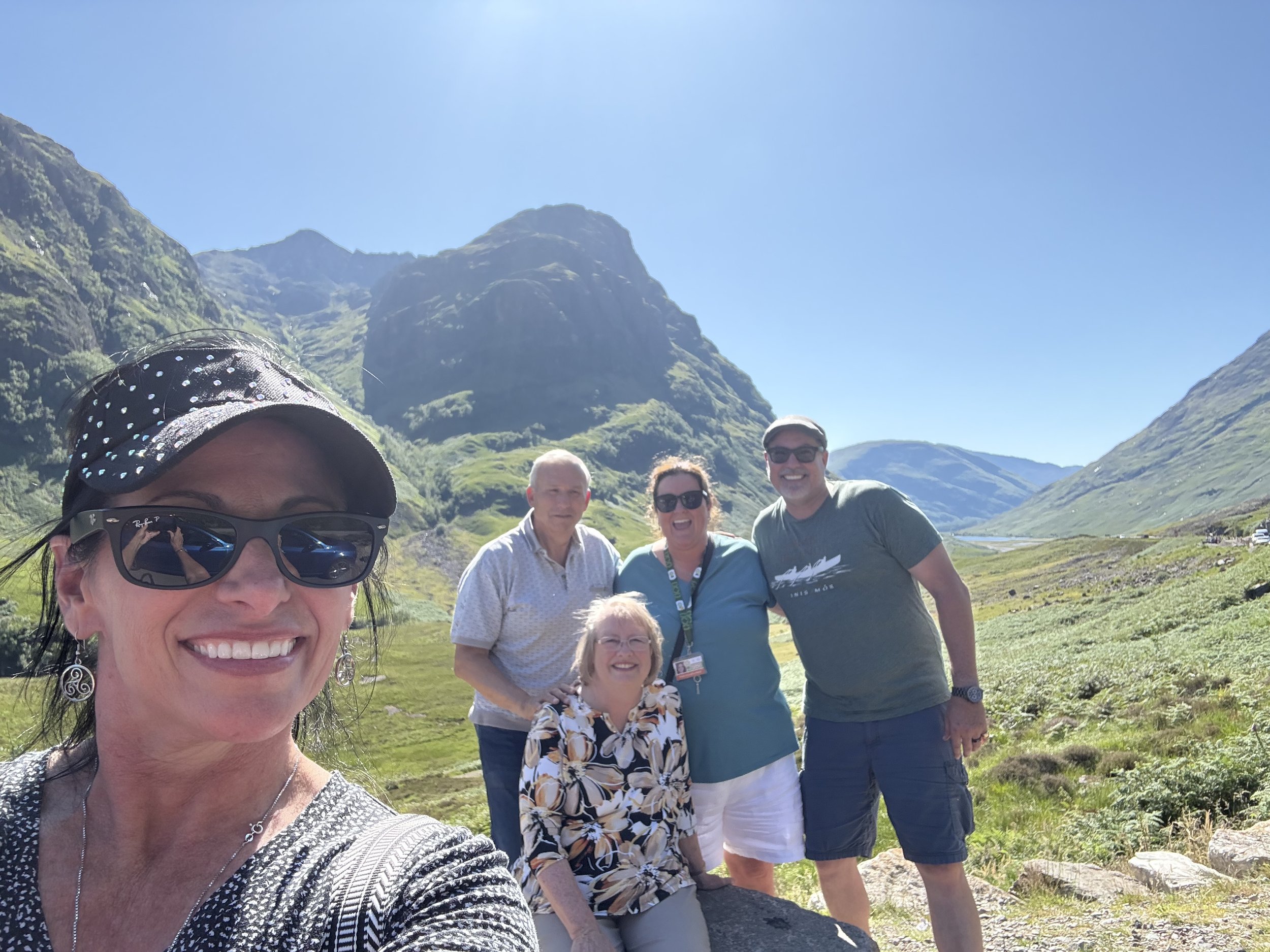 A group of six people smiling and posing outdoors in a mountainous area with green hills and a clear blue sky.