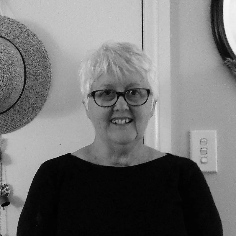 A woman with short, light-colored hair and glasses, smiling, standing indoors near a wall with a round mirror and a light switch.
