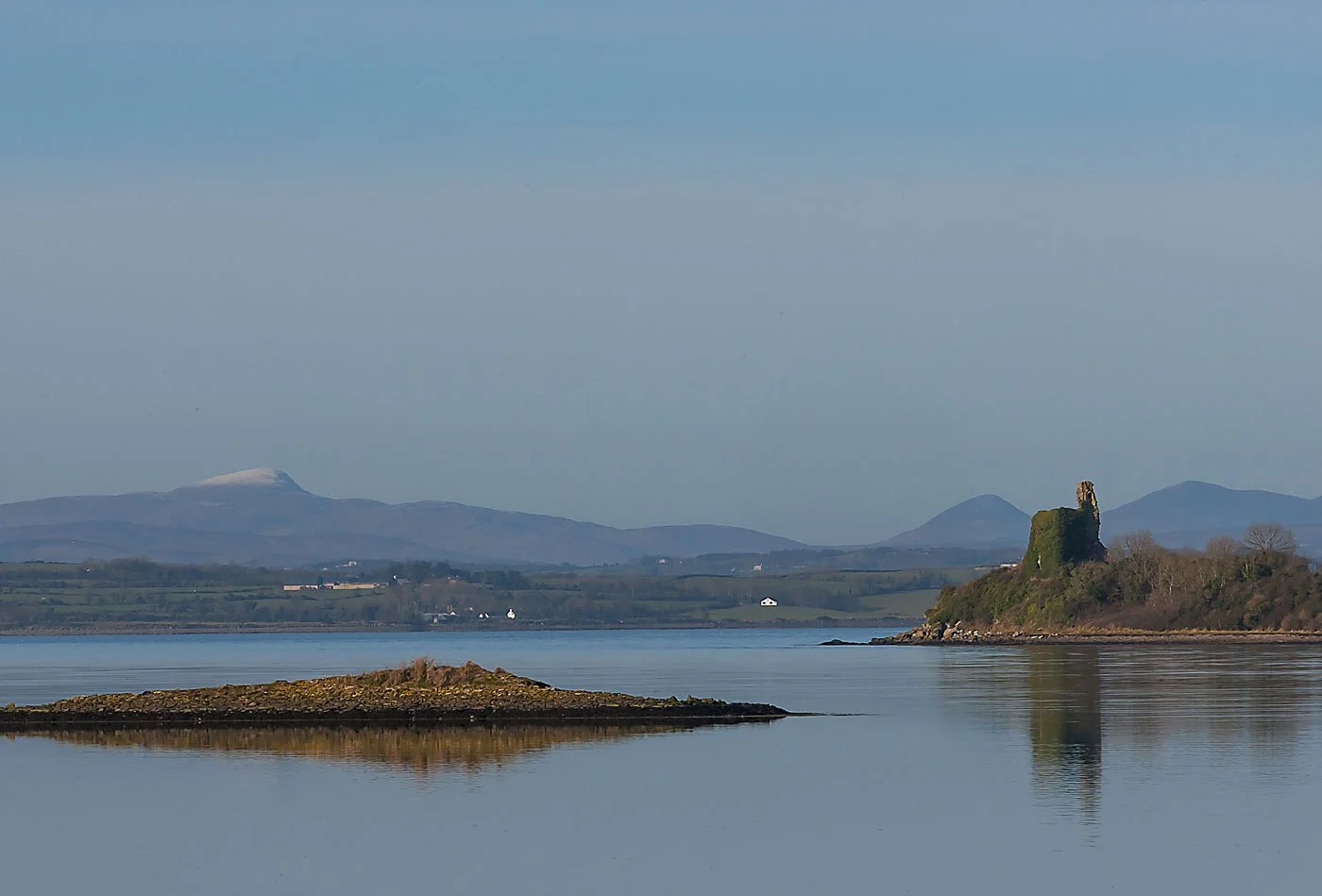 Scenic view of a lake with a small island and an ancient stone tower, with mountains in the background and a clear sky.