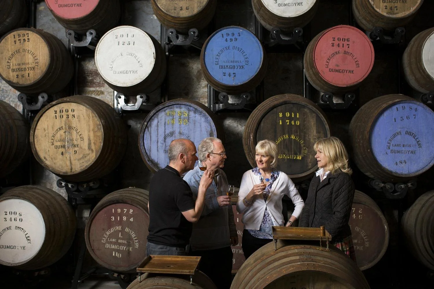 Four people in a wine cellar with large wine barrels in the background, holding glasses of wine and talking.