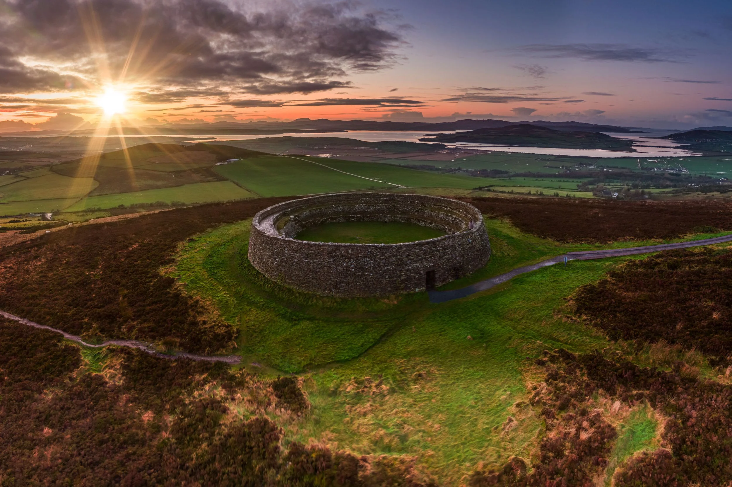 Aerial view of a historic stone fortification on a grassy hillside, overlooking a landscape of fields, lakes, and distant mountains at sunset with a partly cloudy sky.