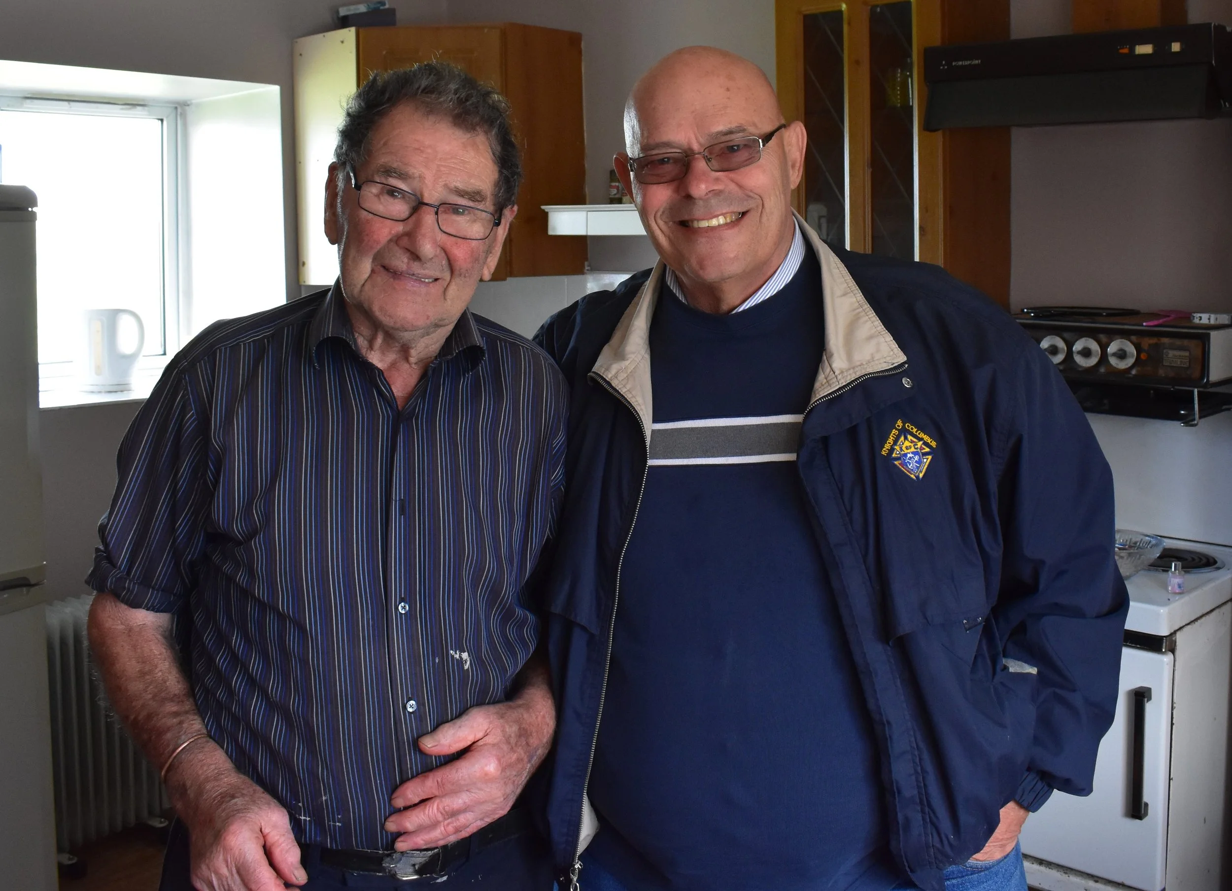Two smiling men standing in a kitchen close to each other, one elderly with glasses and the other middle-aged, both wearing casual clothing.