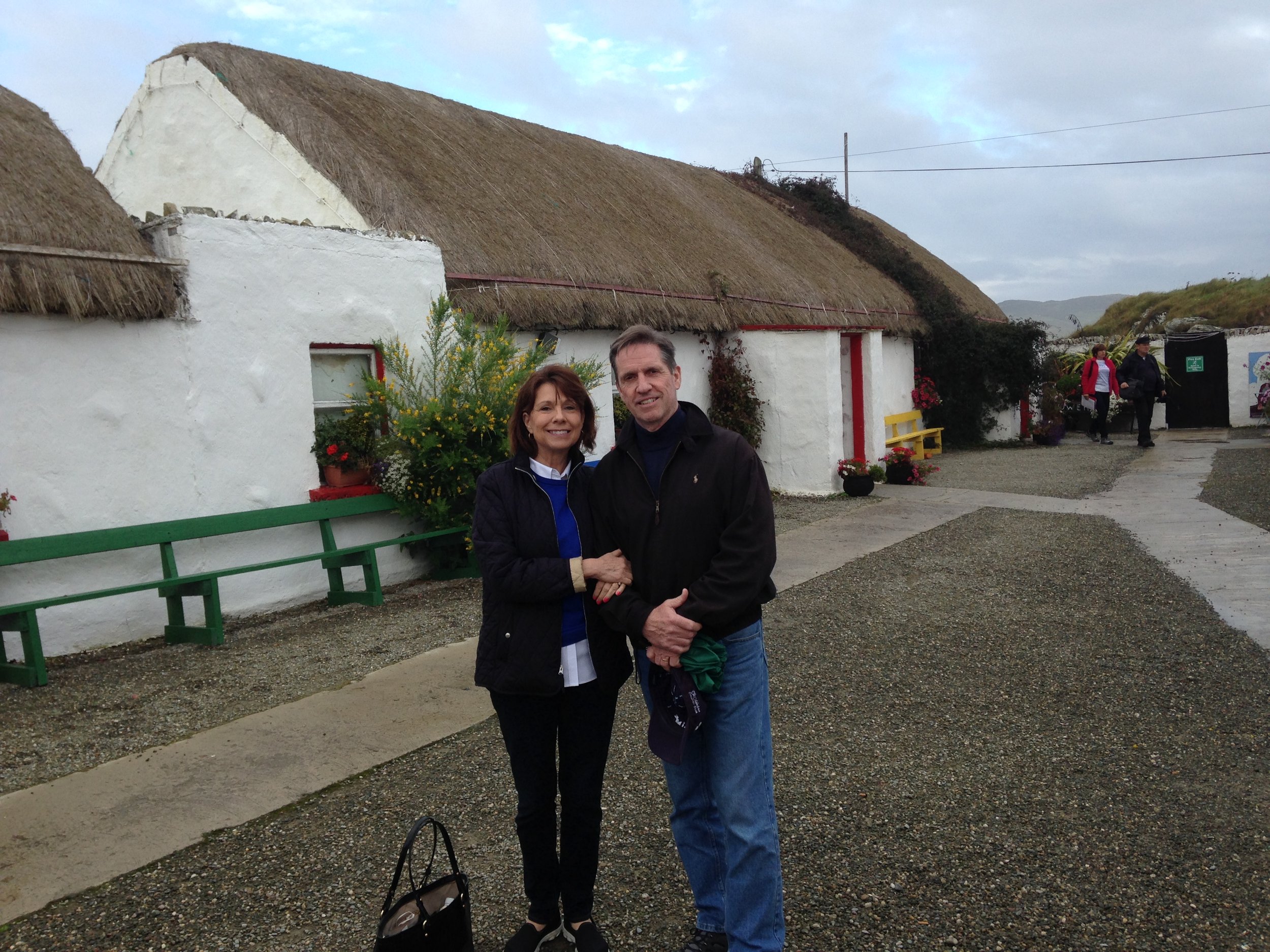 Two people are standing outside a white building with a thatched roof, with a person in a red jacket and another in black walking in the background.