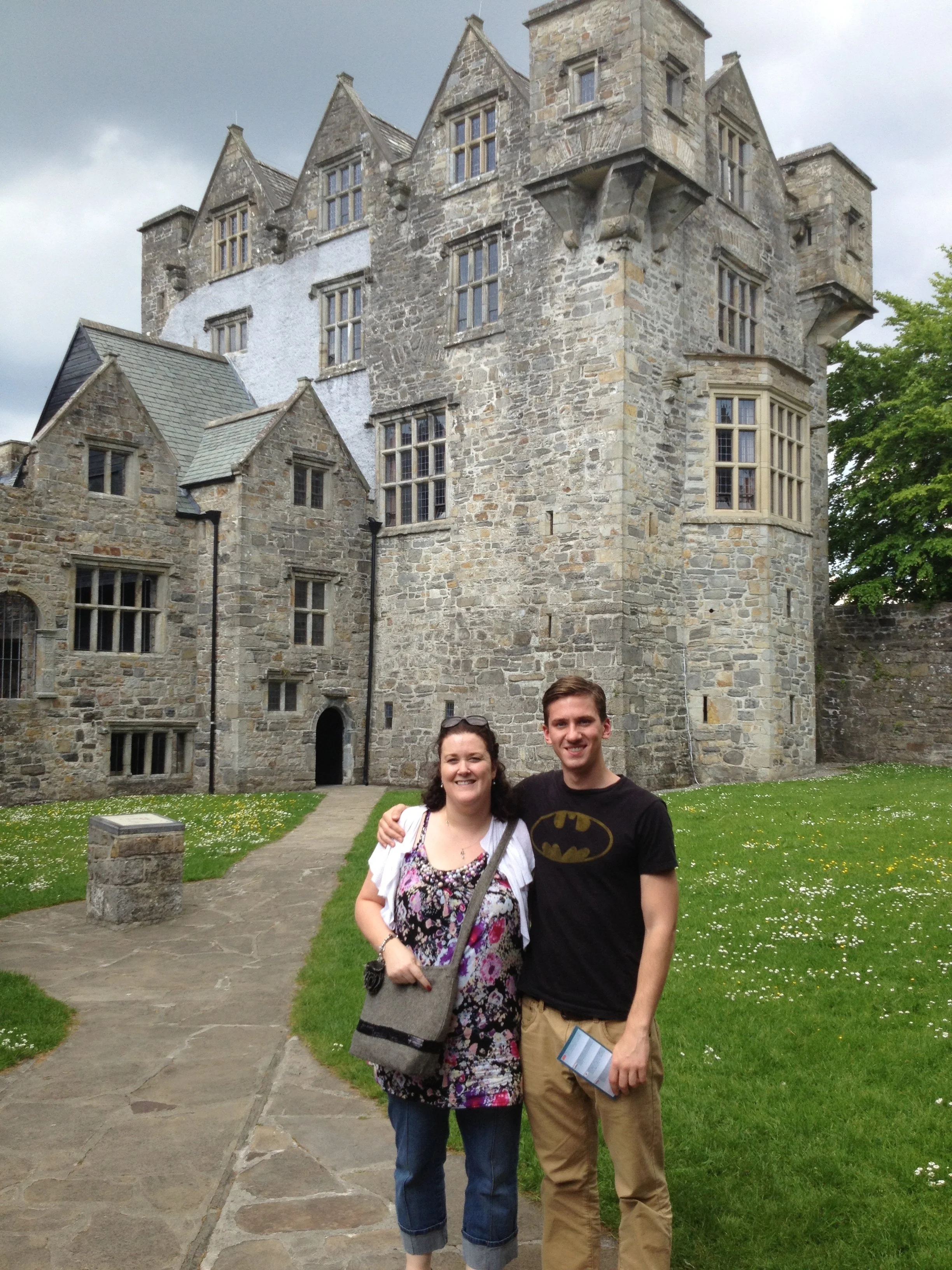 A woman and a man standing outside in front of a stone castle or historic building with multiple turrets and windows. The woman is wearing a floral dress, carrying a bag, and has sunglasses on her head. The man is wearing a Batman t-shirt and khaki pants, holding a brochure or pamphlet. The sky is cloudy, and there is a patch of grass with small flowers around them.