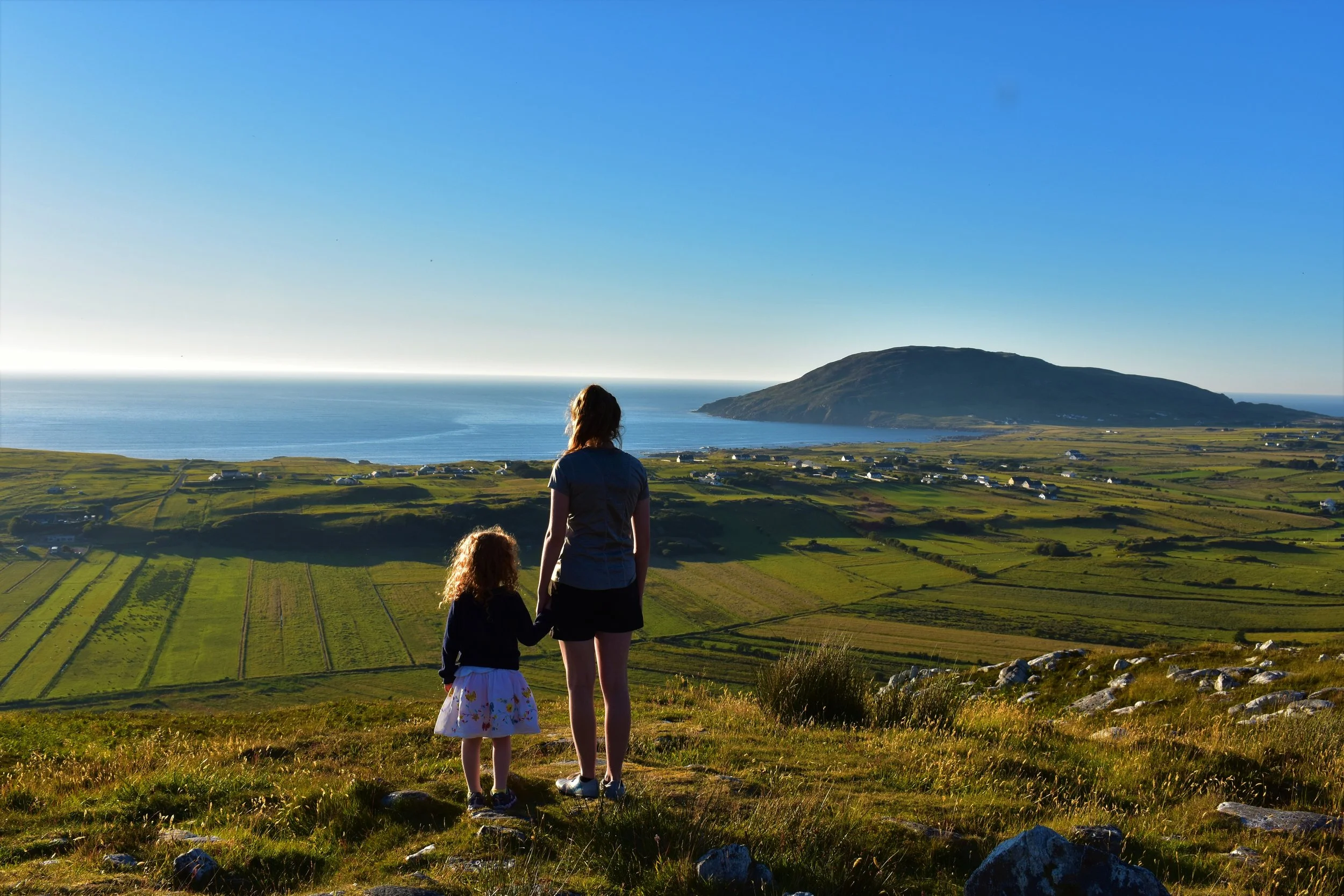 Three people, a woman and two children, standing on a grassy hill overlooking a scenic landscape of green fields, a large hill or mountain, the ocean, and a clear blue sky.
