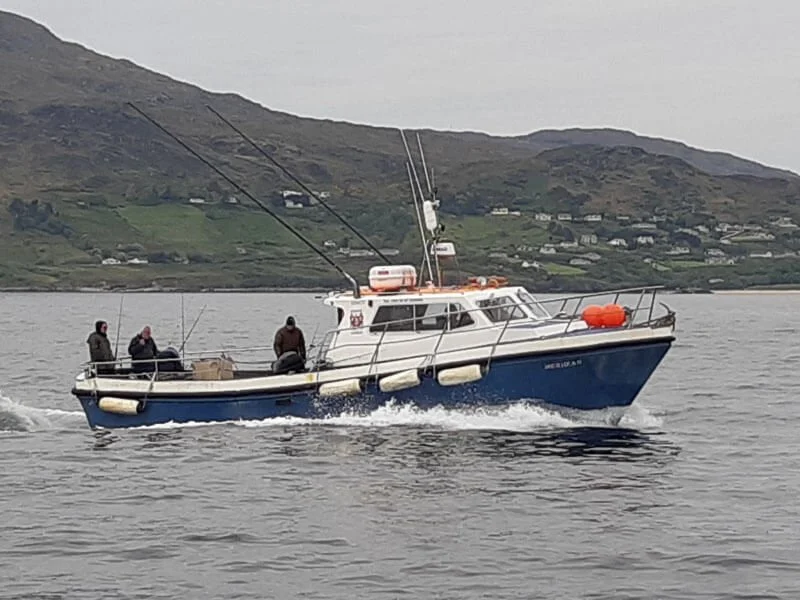 A white and blue fishing boat sailing on a body of water with three people on board, fishing rods, orange flotation devices, and a mountainous landscape in the background.