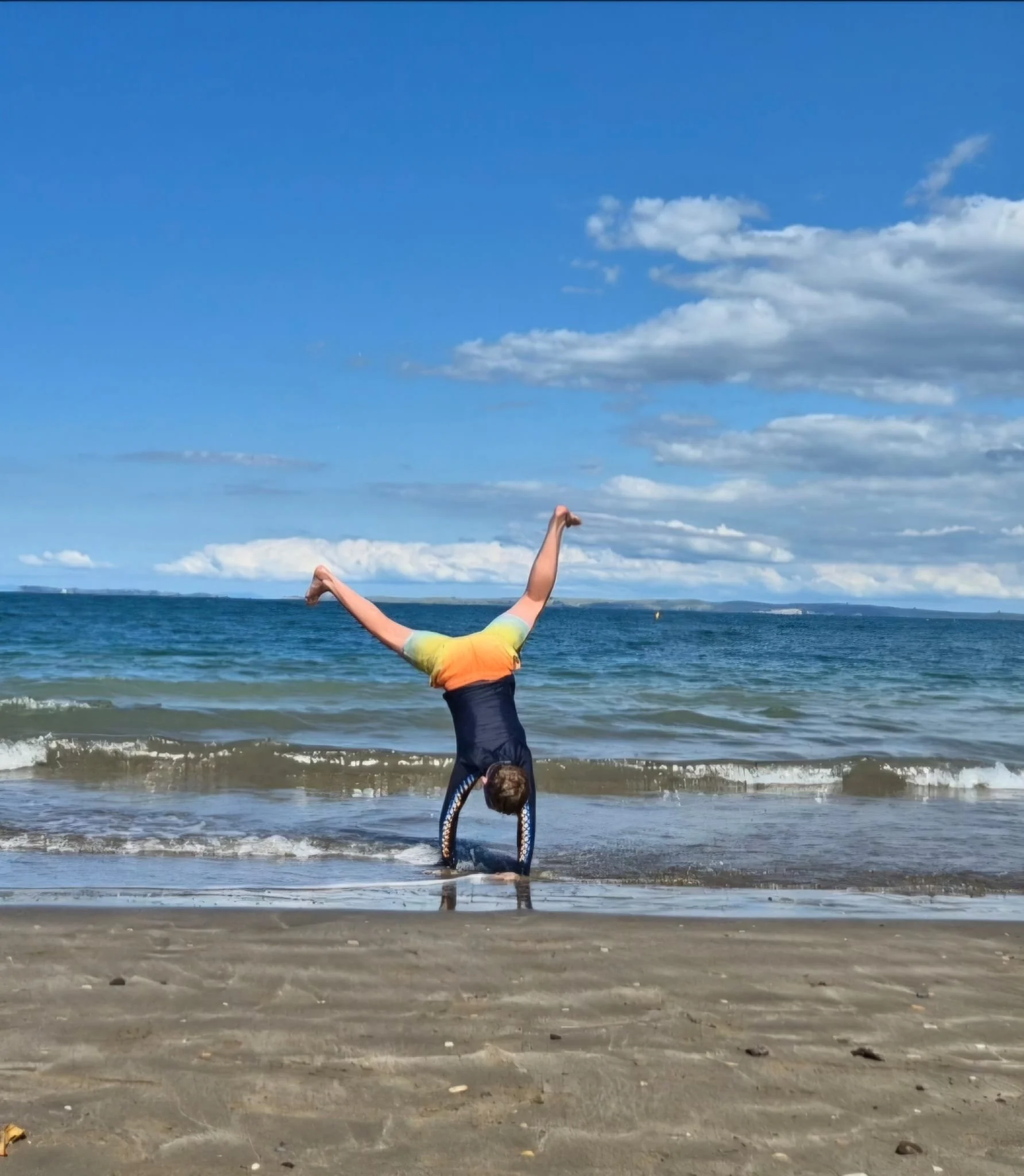 Person performing a handstand on the beach near the shoreline with the ocean and blue sky with clouds in the background.