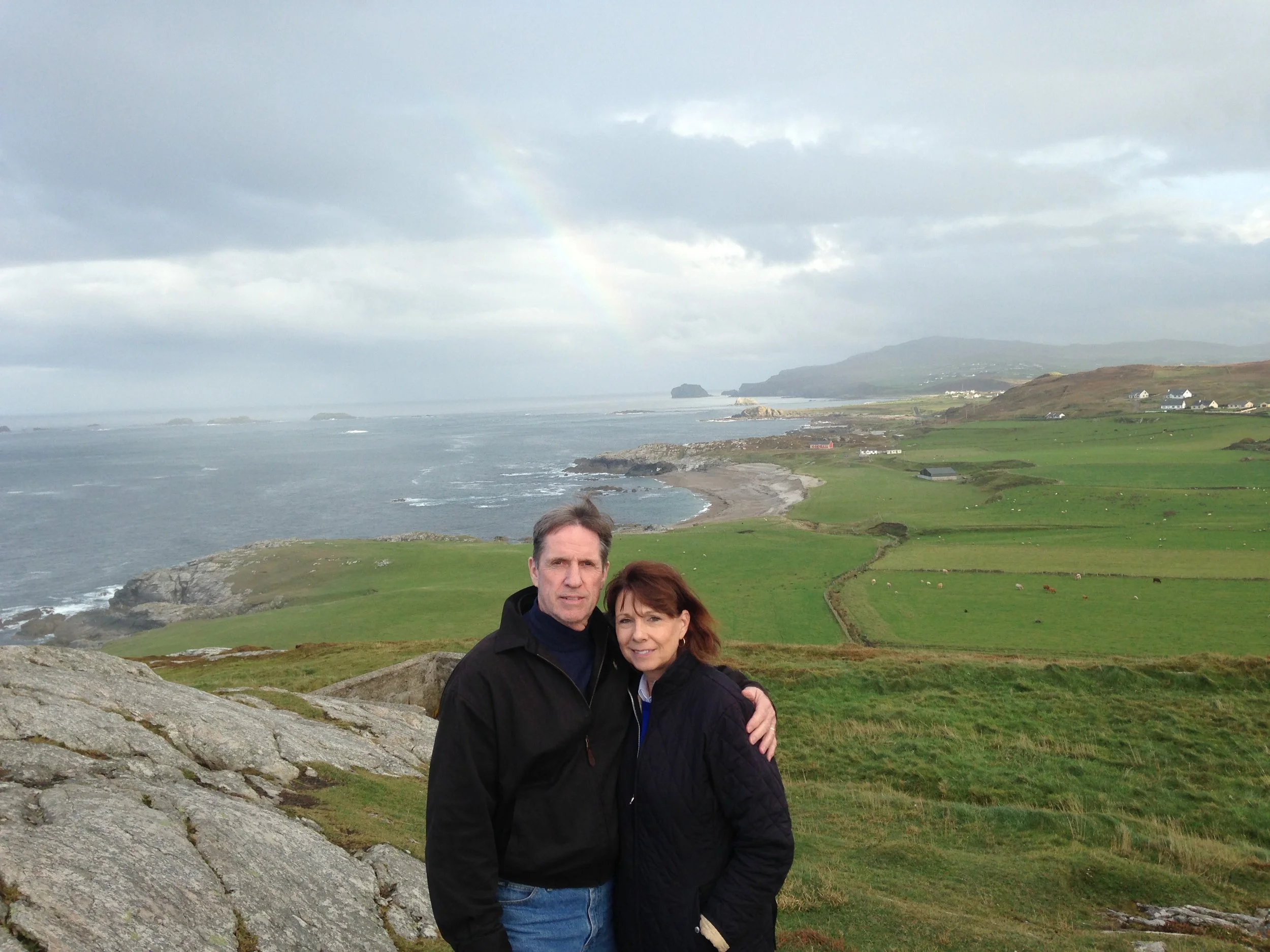 A couple standing outdoors on a grassy and rocky area near the coast, with a cloudy sky, a faint rainbow, and a distant shoreline with houses and hills in the background.