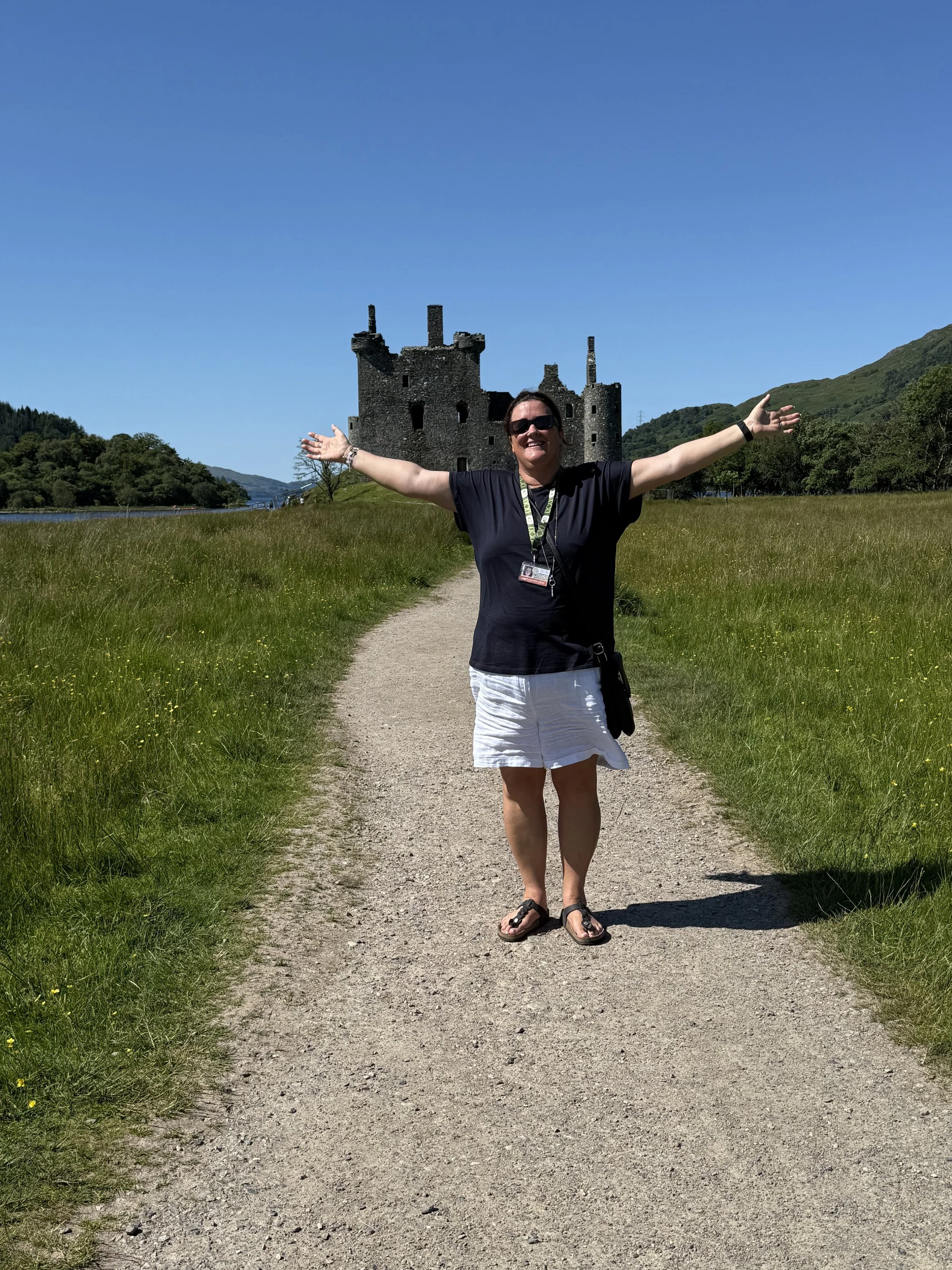 Woman smiling with open arms standing on a dirt path in front of a historic castle on a sunny day.