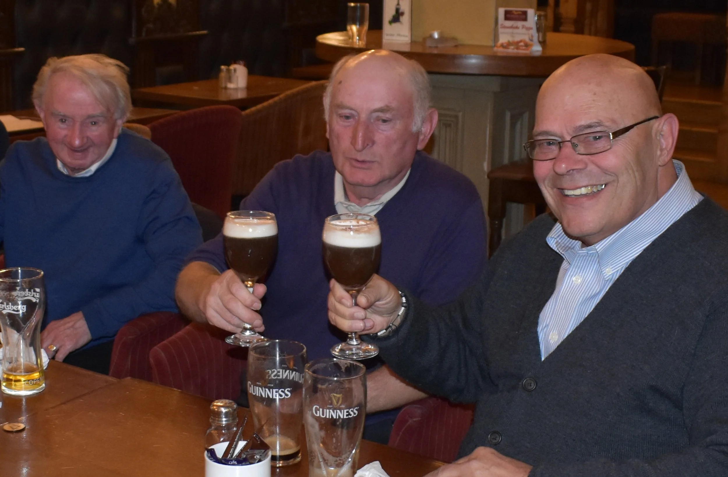 Three men sitting at a bar or restaurant table, each holding a glass of dark beer with a foamy head, smiling and celebrating together. The table has glasses and some eating utensils. The background shows a bar or pub setting with dim lighting.