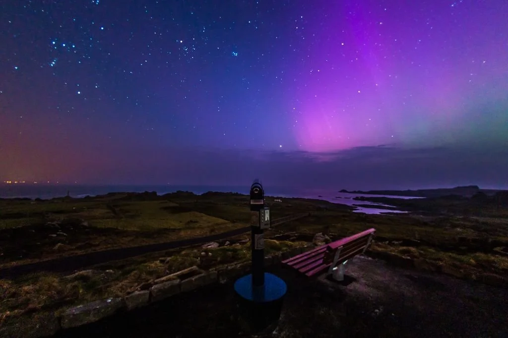 A bench on a hill overlooks the ocean at night with a colorful aurora borealis and stars in the sky.