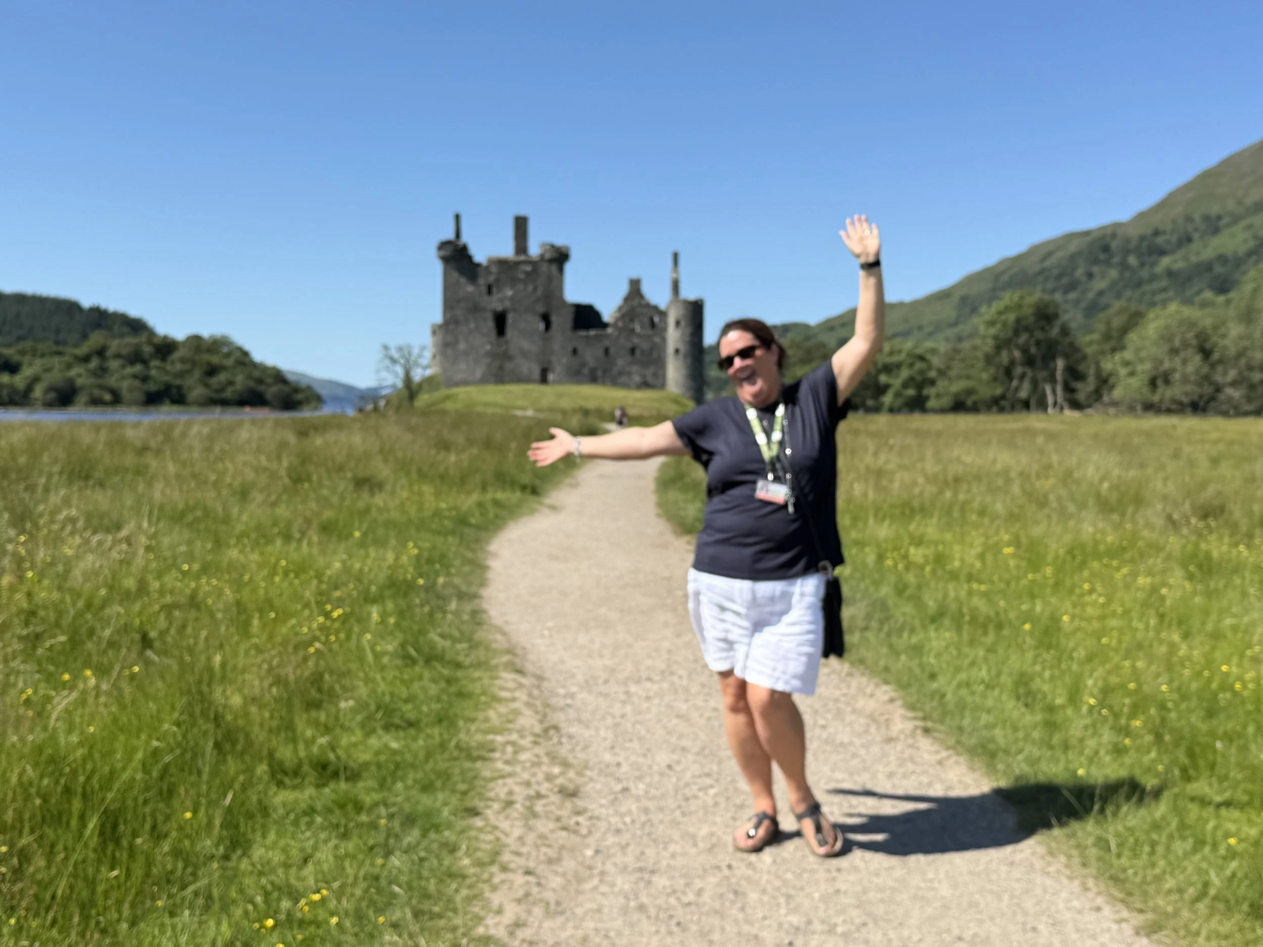 A woman wearing sunglasses, a navy blue t-shirt, white shorts, and sandals standing on a dirt path with her arms outstretched in front of a castle surrounded by green hills and a blue sky.