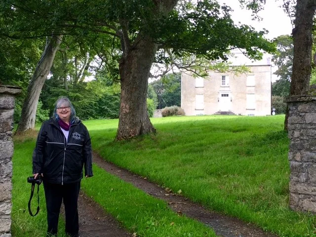 Woman standing on a grass path in a park with trees and an old stone building in the background.