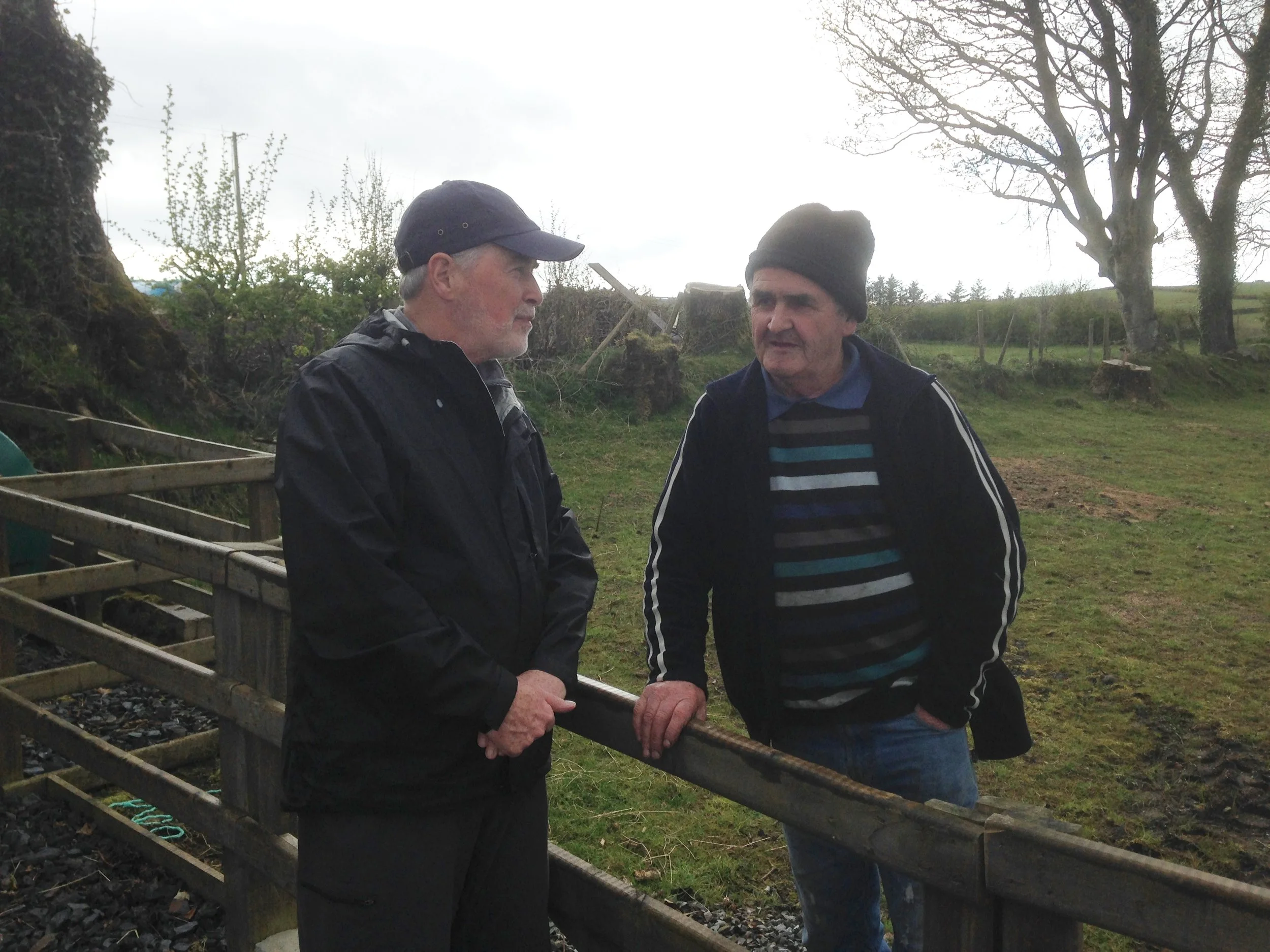 Two older men are outside near a small train track, standing on either side of a guardrail, engaged in conversation. One is wearing a dark jacket and cap, and the other is in a striped sweater and black beanie. Trees and open fields are in the background under an overcast sky.