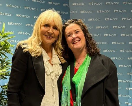 Two women smiling in front of a backdrop with RTÉ Radio 1 logos.