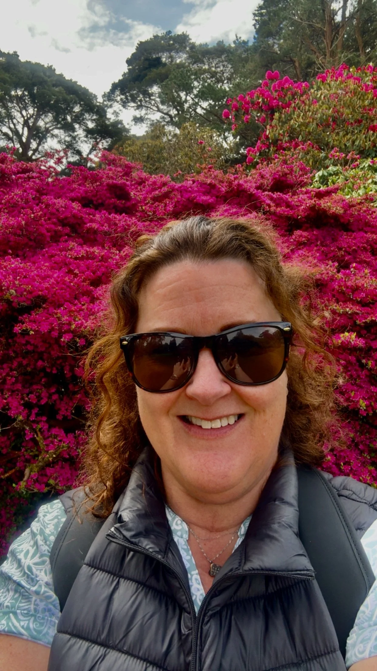 A woman with curly reddish hair and sunglasses smiling in front of bright pink flowers and green trees.