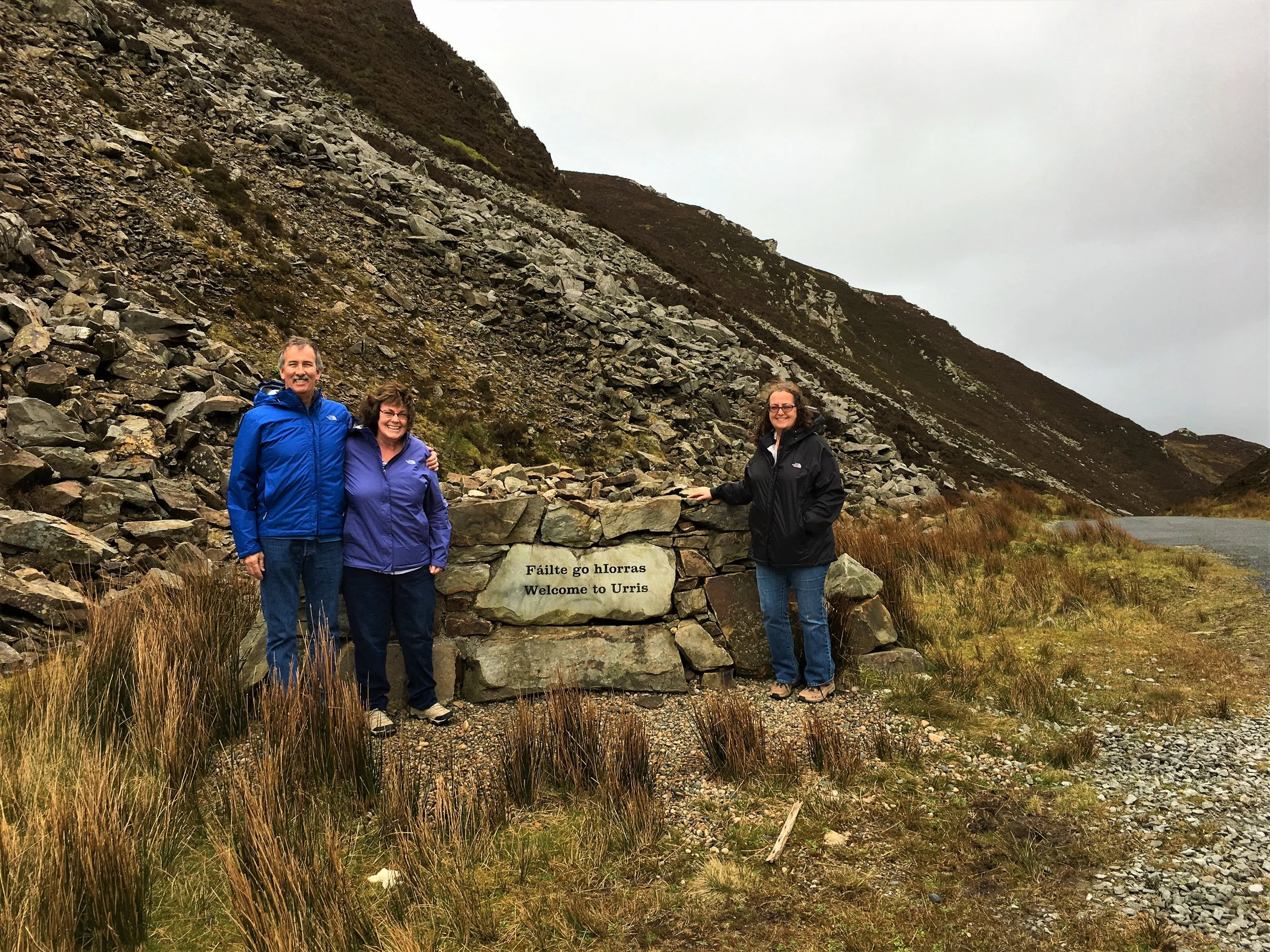 Three people standing outdoors in front of a stone marker that says "Fáilte go hIorras Welcome to Urris". They are dressed in outdoor jackets, and there is a rocky hillside and an overcast sky in the background.