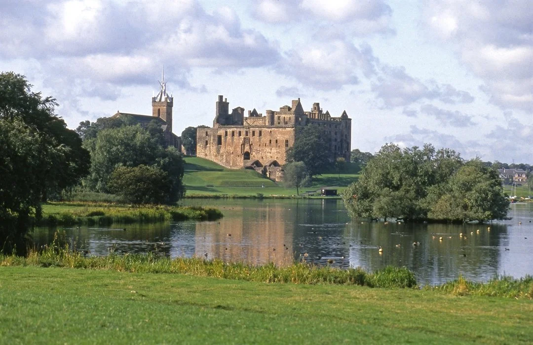 A historic stone castle located near a body of water, with lush greenery and trees in the foreground, and cloudy skies overhead.