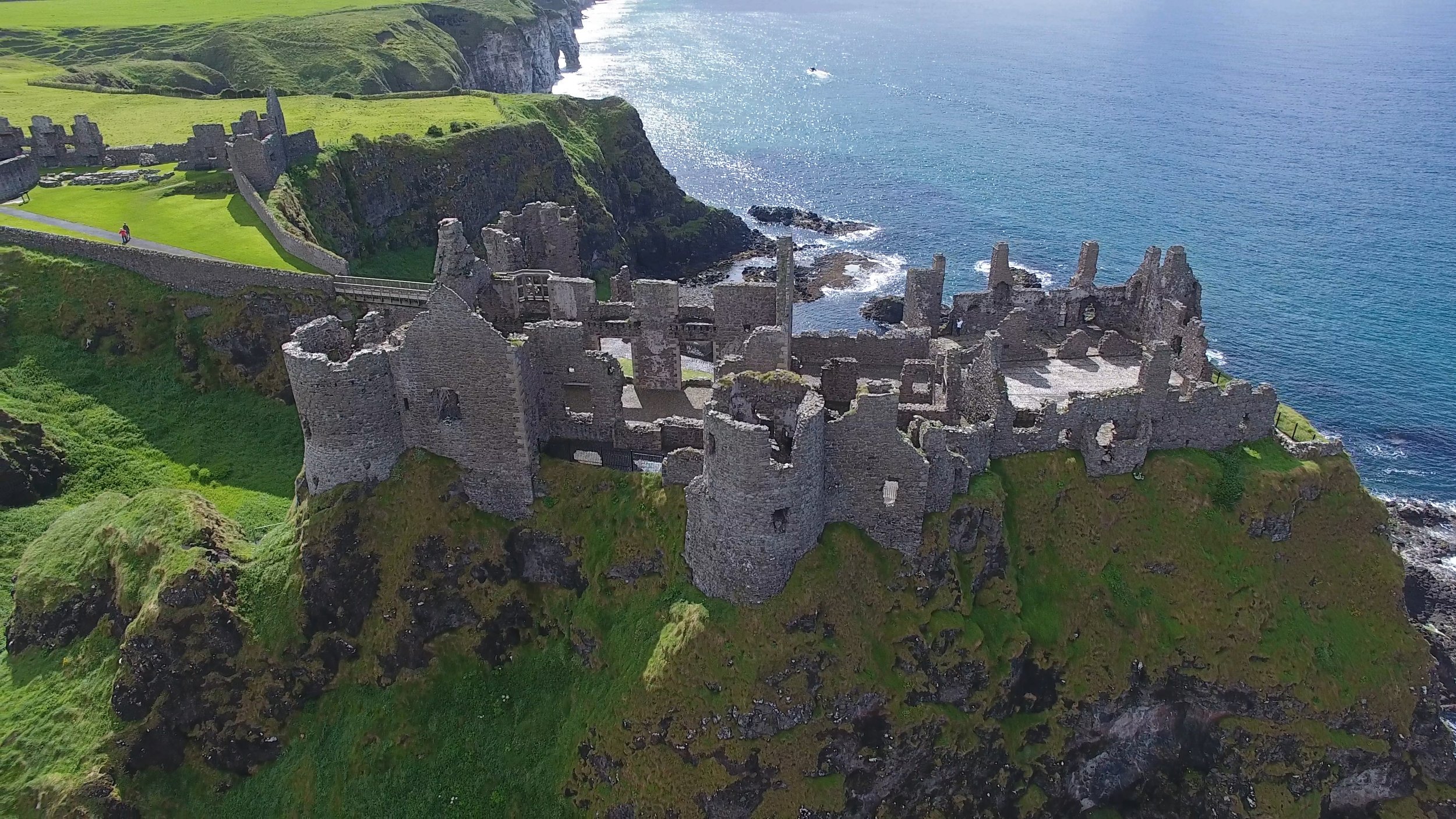 Ruins of an ancient stone castle on a cliff overlooking the ocean, with green grassy areas and a pathway, under a clear sky.