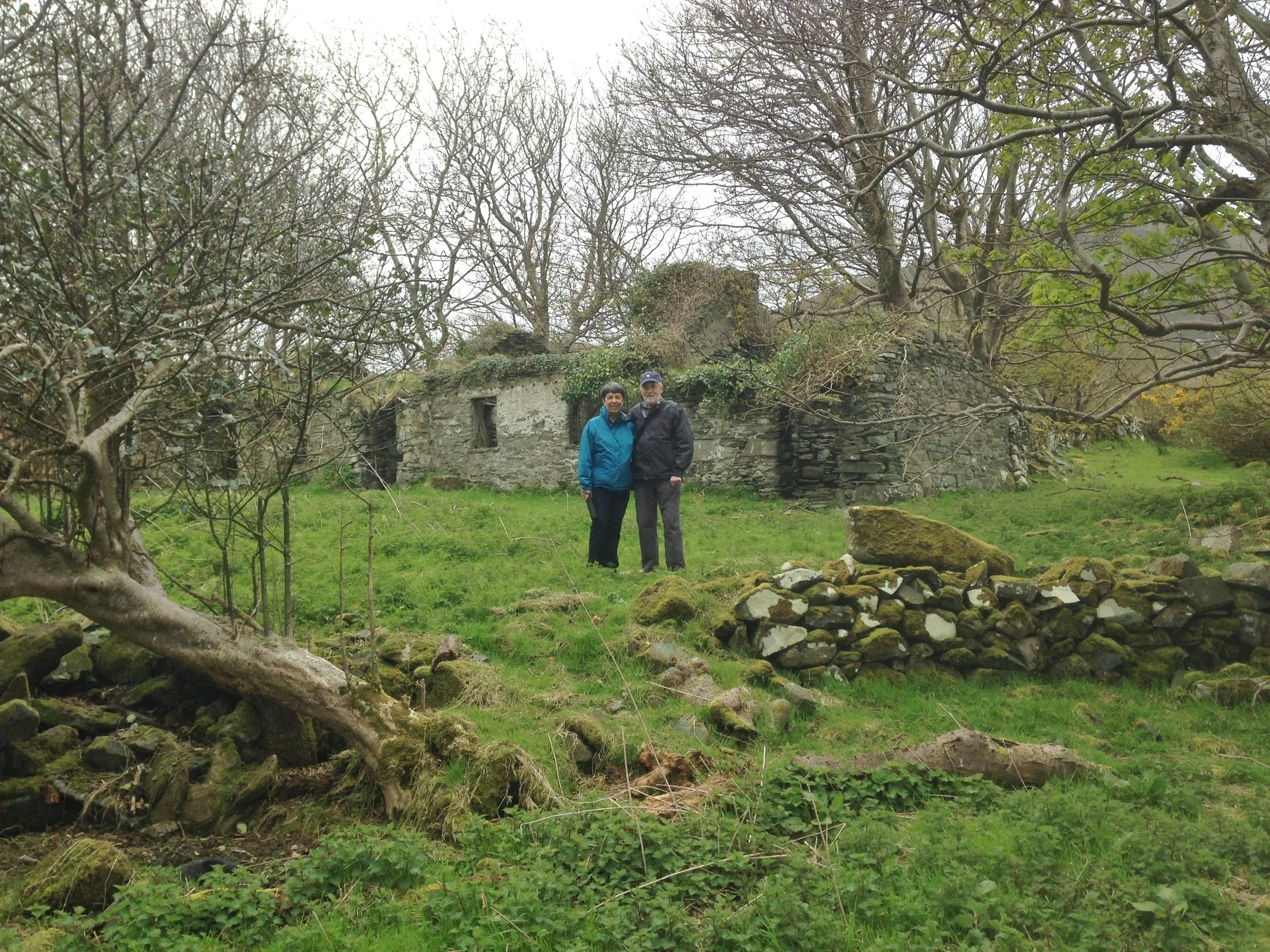 Two people standing together outdoors in front of an old stone structure surrounded by leafless trees and green grass.