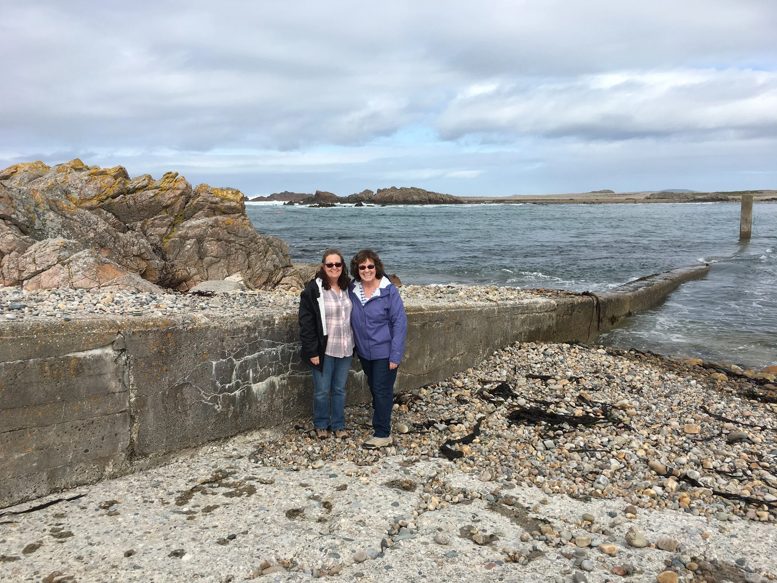 Two women smiling and standing near a rocky shoreline with water and clouds in the background.